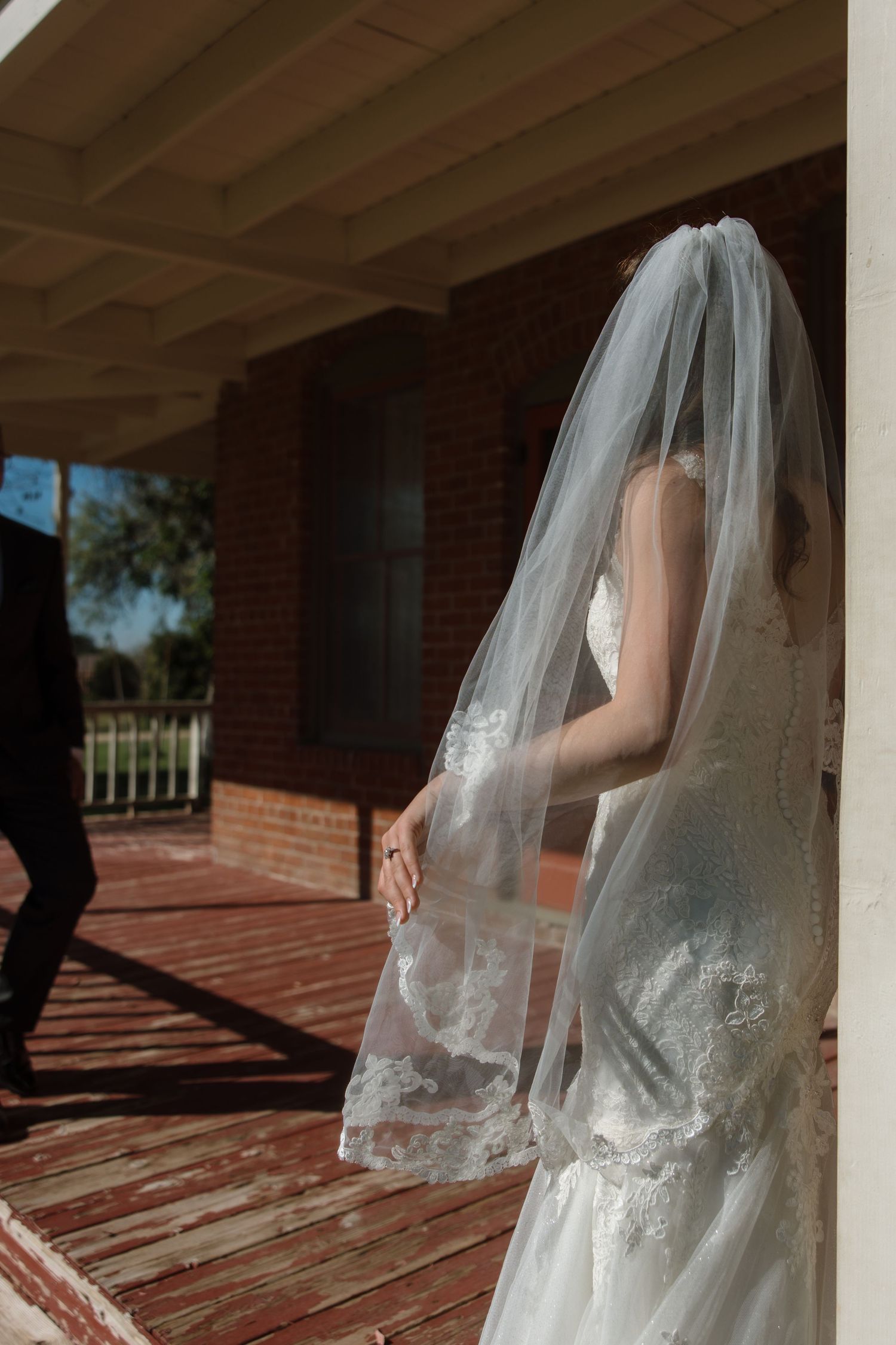 Bride in lace wedding gown and veil stands on a sunlit porch as groom approaches from behind.