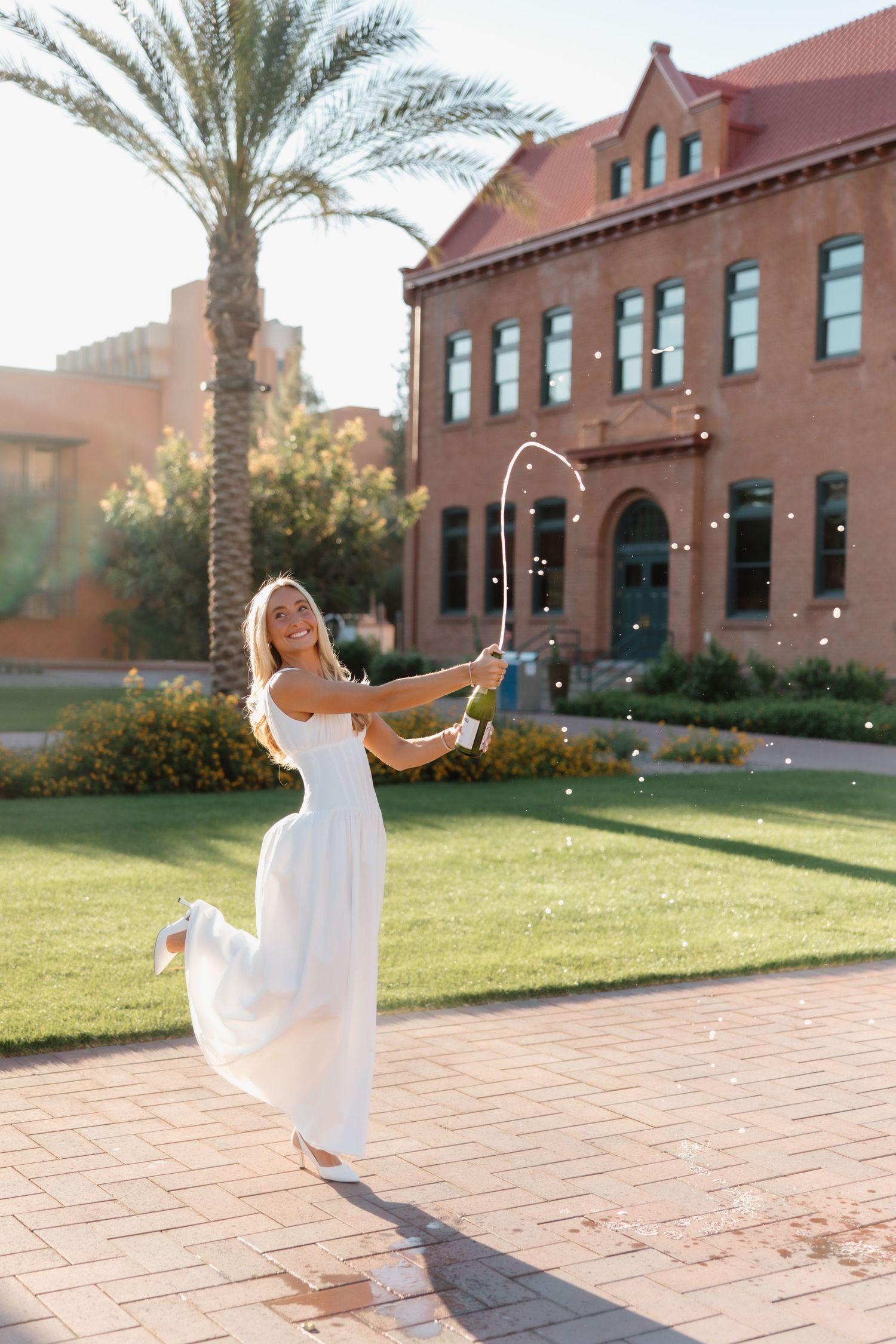A joyful woman in a white dress pops champagne outdoors near a red brick building and palm tree.
