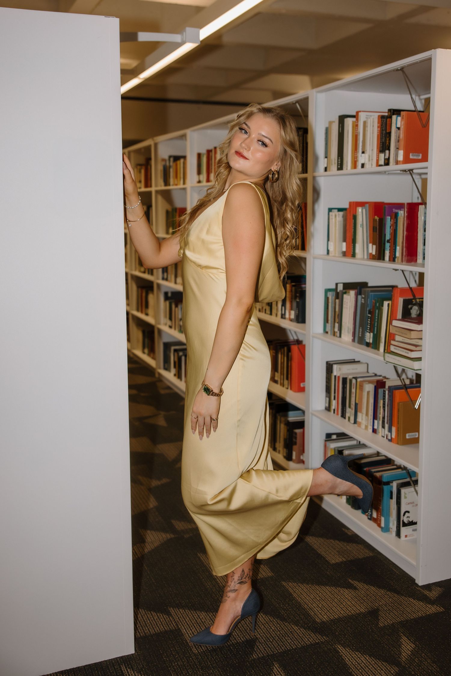 Woman in gold satin dress posing elegantly between library bookshelves wearing blue heels.