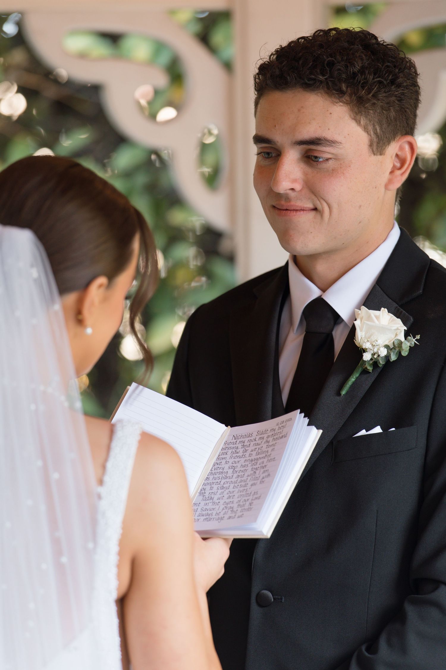 Smiling groom listens as bride reads vows from notebook at elegant outdoor wedding ceremony.