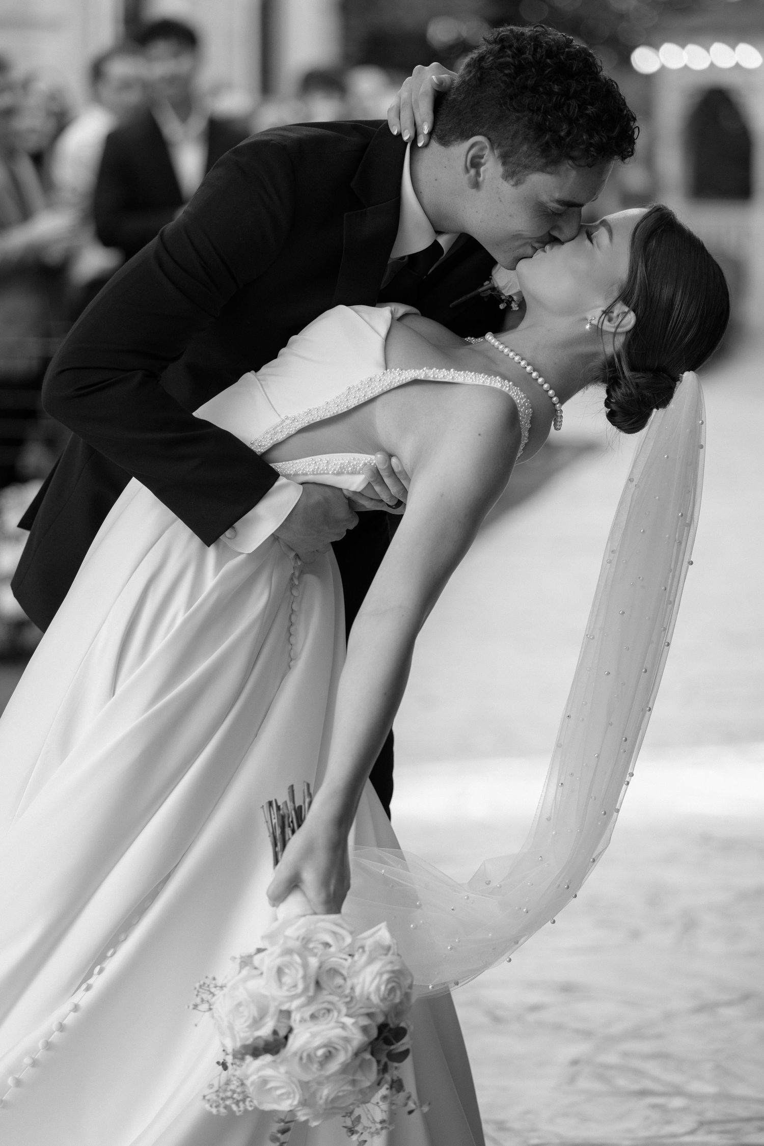 Black and white photo of groom dipping bride for passionate kiss holding white rose bouquet.
