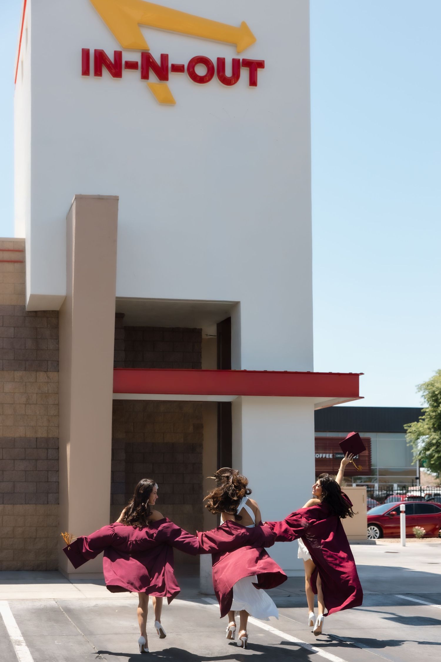 Three graduates in maroon gowns running toward In-N-Out Burger on a sunny day.