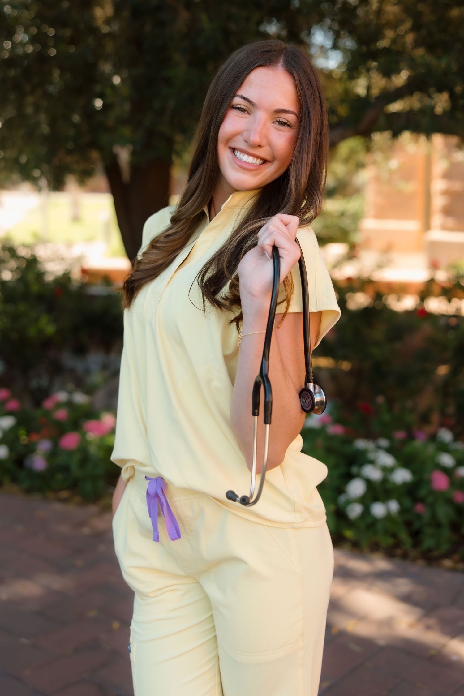 Smiling female nursing graduate in yellow scrubs holding stethoscope in blooming campus garden.