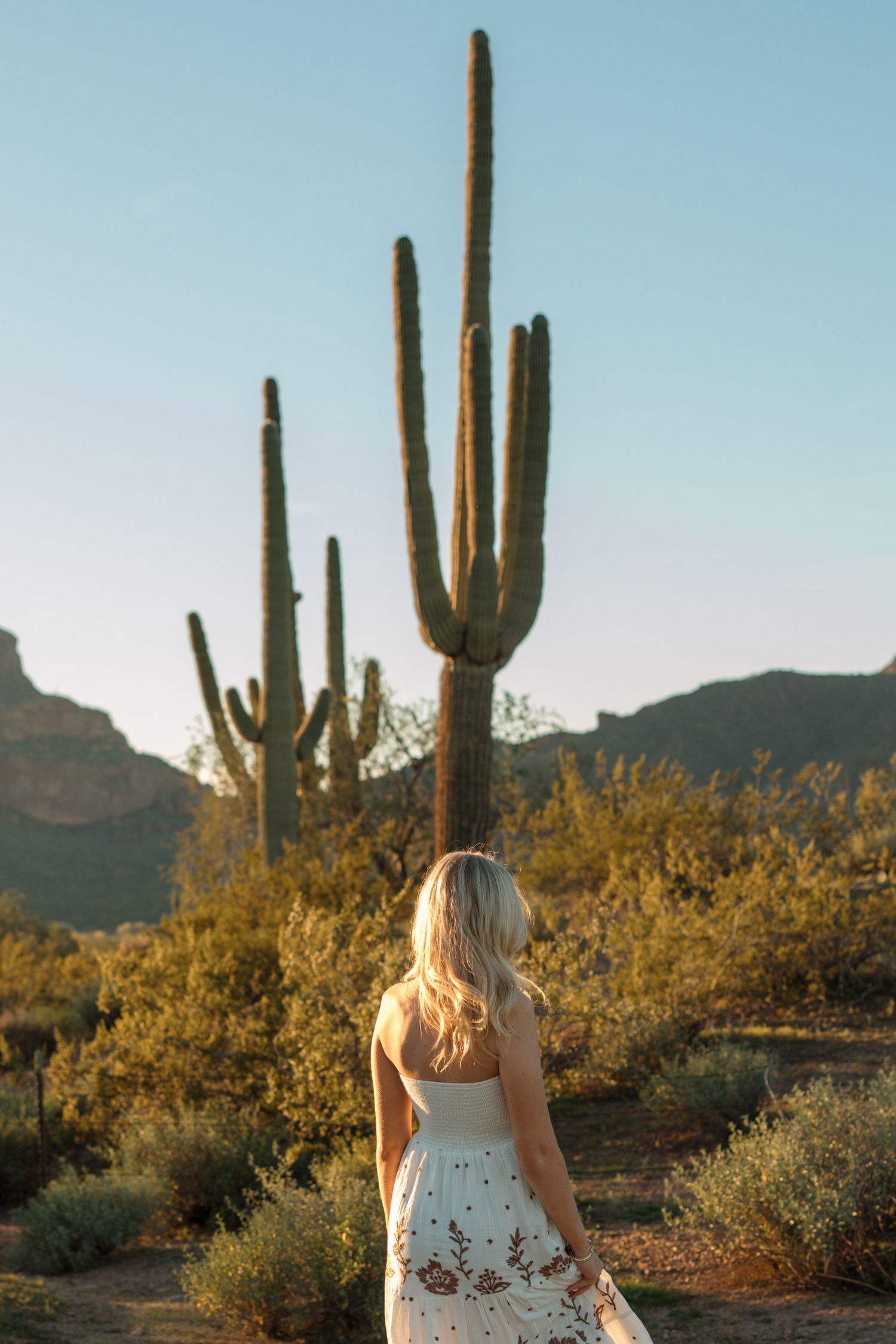 Blonde woman in white embroidered maxi dress walking toward tall saguaro cacti in Arizona desert landscape.