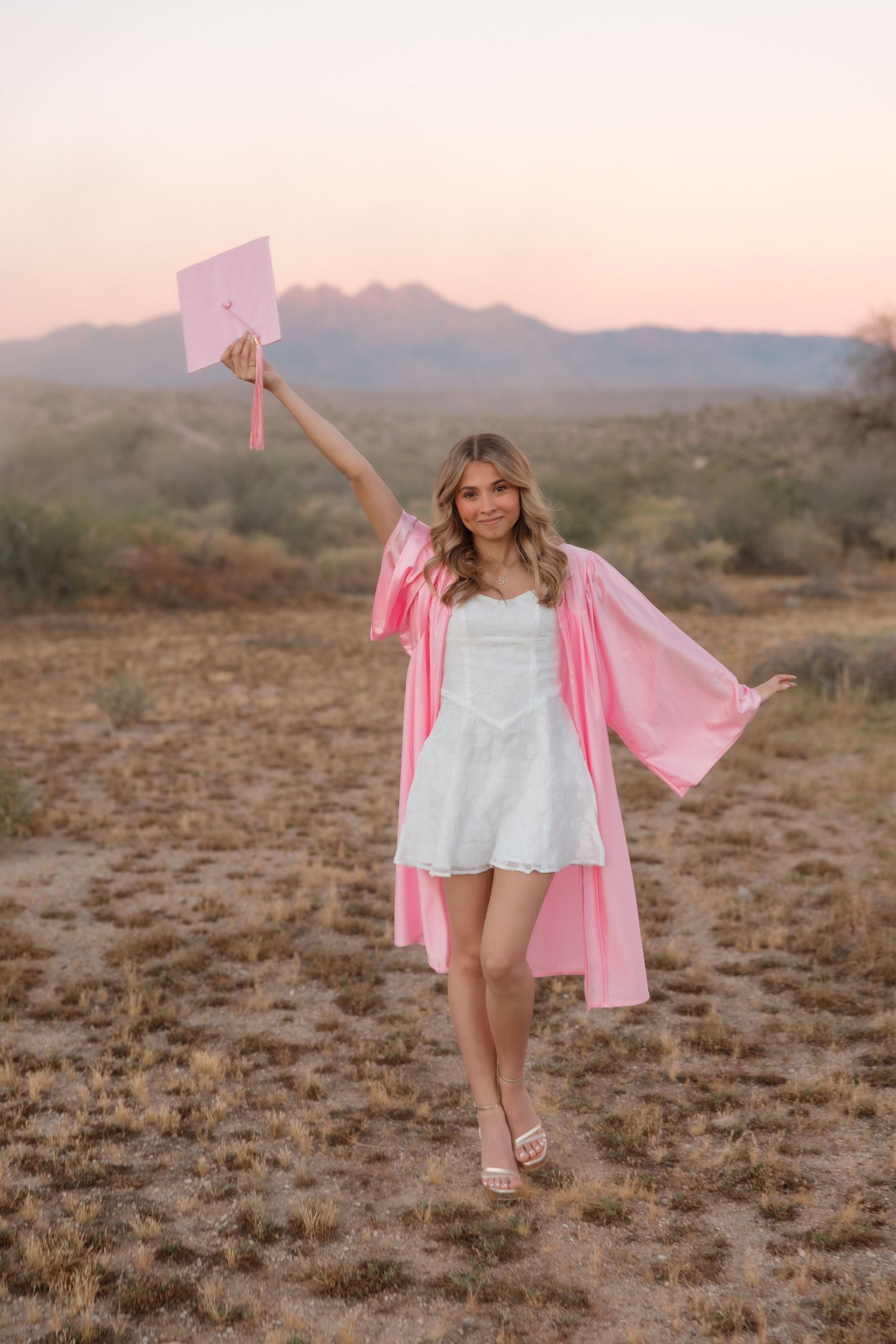Young woman celebrating graduation in pink cap and gown raising cap in desert landscape at sunset.