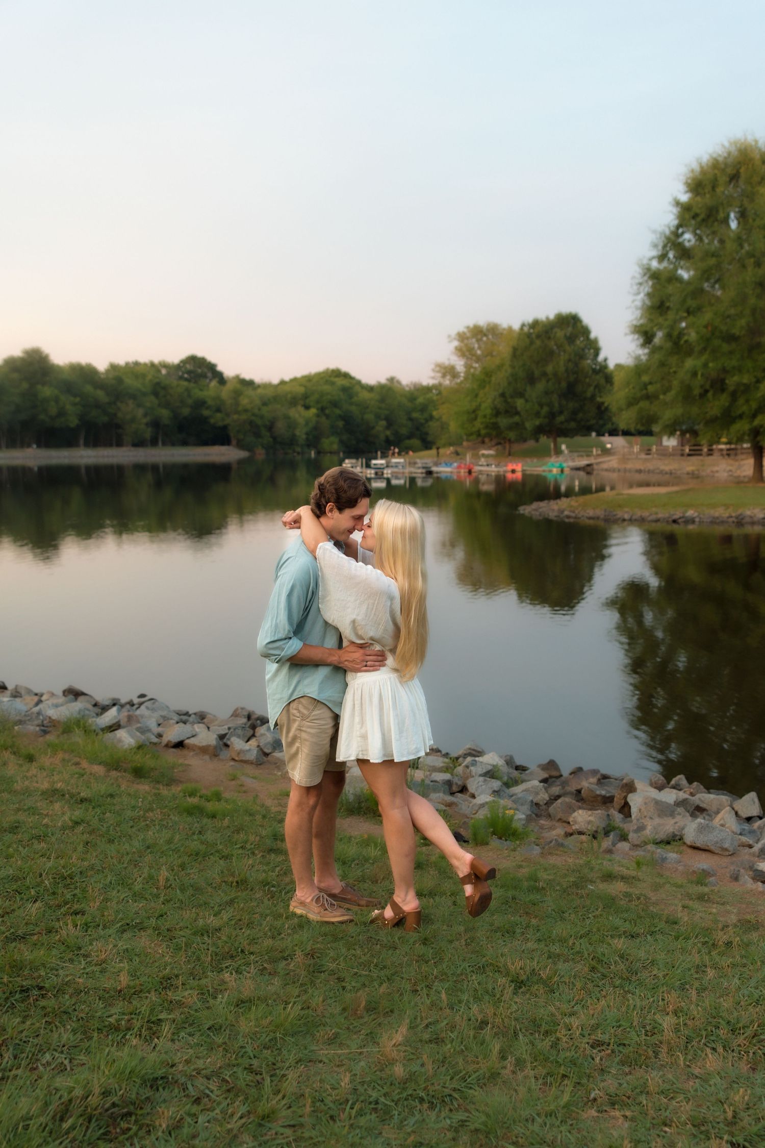 Romantic couple sharing a kiss by a peaceful lake at dusk in a scenic outdoor setting.