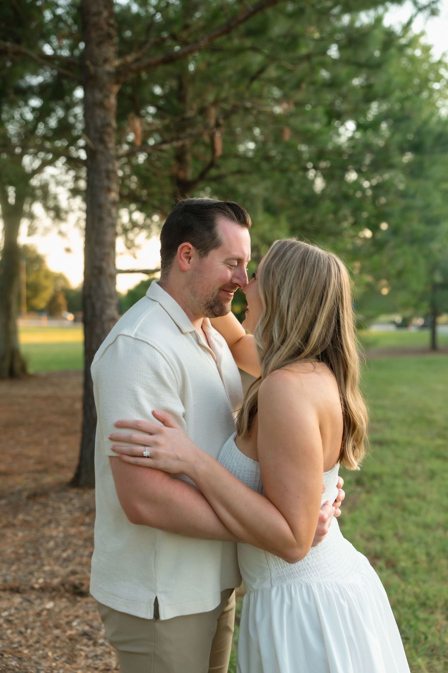 Couple sharing a romantic almost-kiss embrace outdoors among tall trees during golden hour engagement photo session.