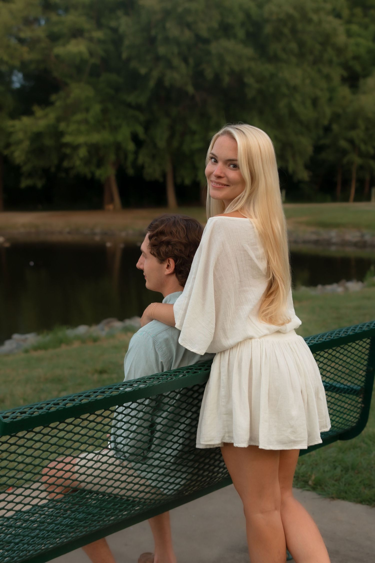 Young blonde woman in white dress smiling at camera while leaning on park bench beside her partner.