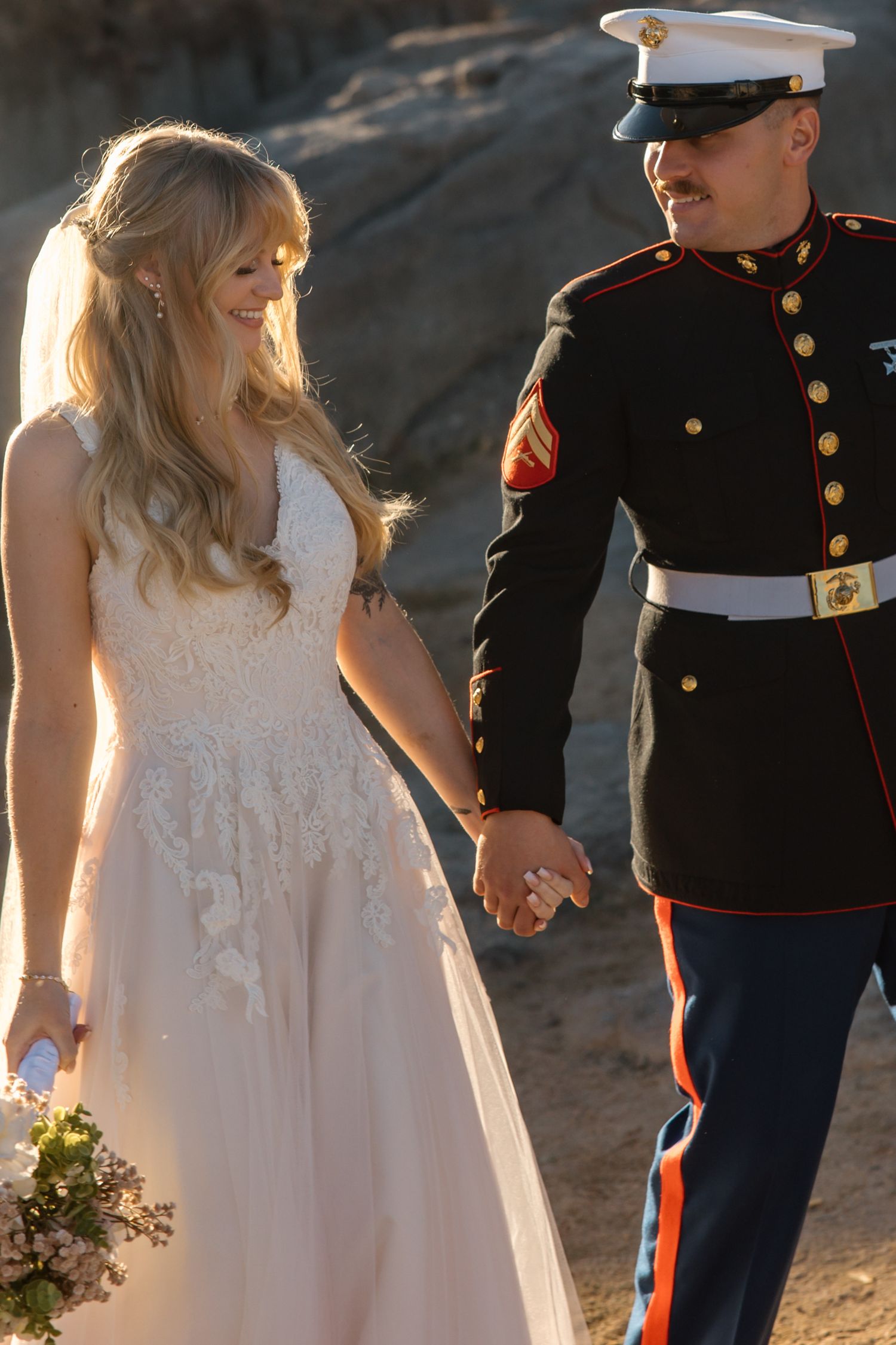 Marine groom in dress uniform holds hands with smiling blonde bride in lace wedding gown outdoors.