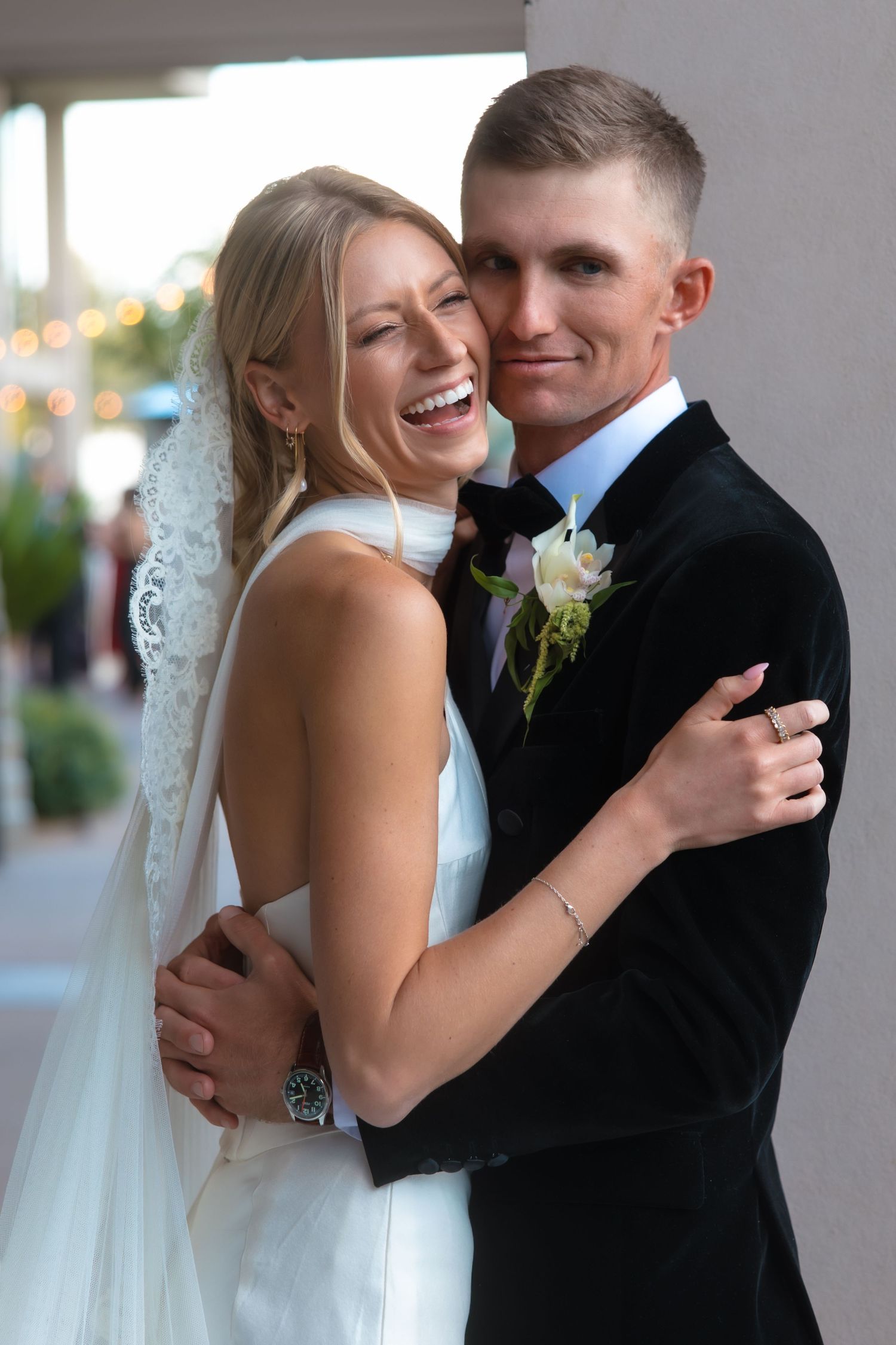 Laughing bride in white halter gown and veil embraces groom in black velvet tuxedo outdoors.