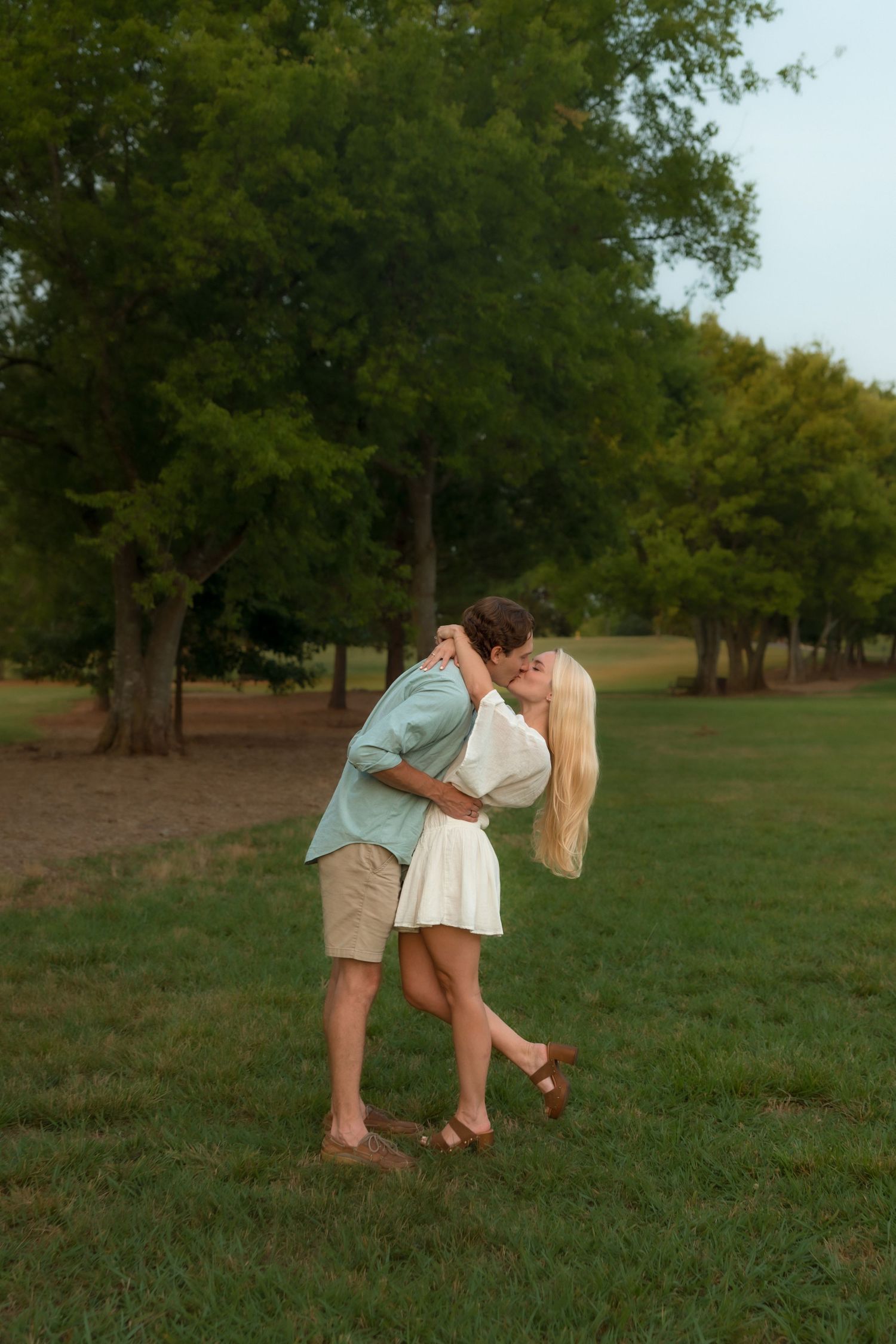 Couple sharing a passionate dip kiss on a grassy field surrounded by tall green trees at dusk.