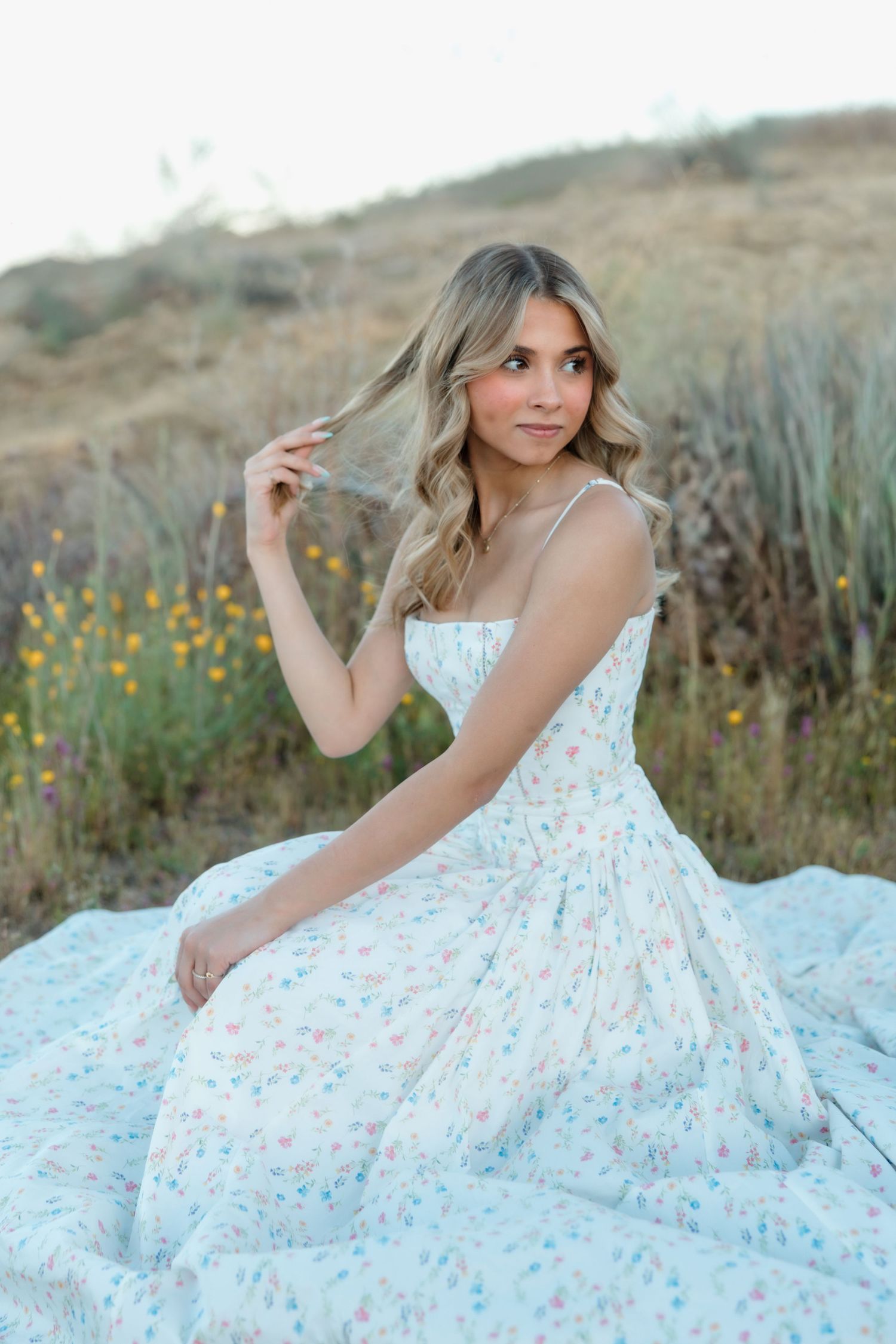 Blonde woman in white floral dress sitting outdoors in field, twirling hair during senior photo session.