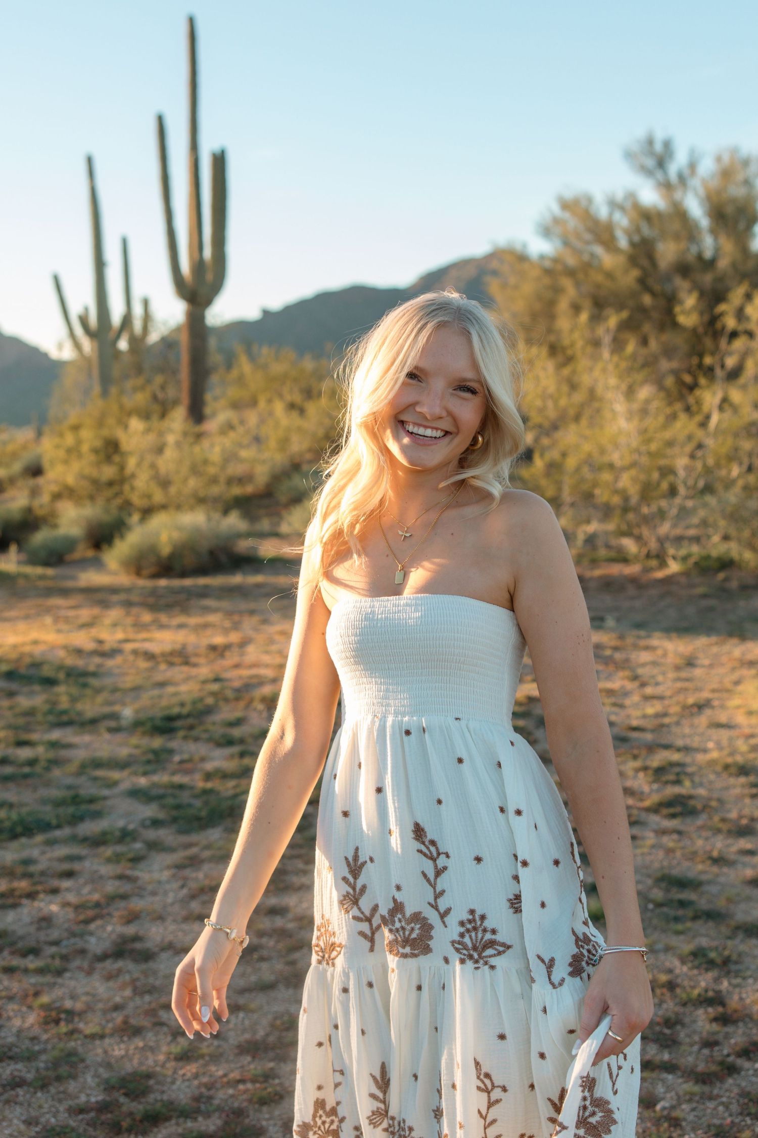 Smiling blonde woman in white floral maxi dress standing in Arizona desert with saguaro cactus at golden hour.