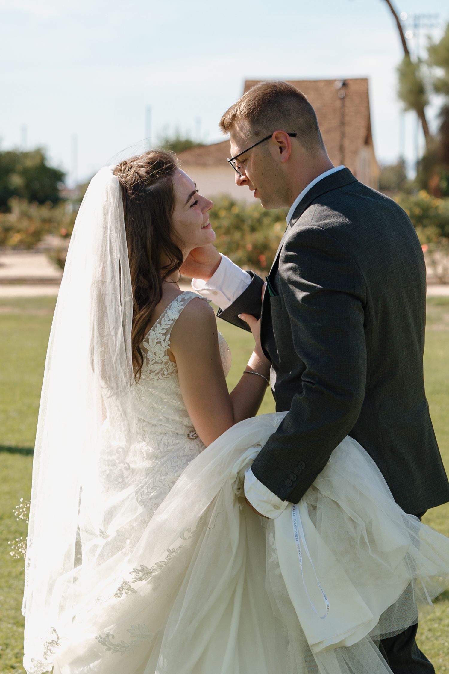 Bride and groom share a tender moment outdoors, with groom gently touching bride's face in wedding attire.