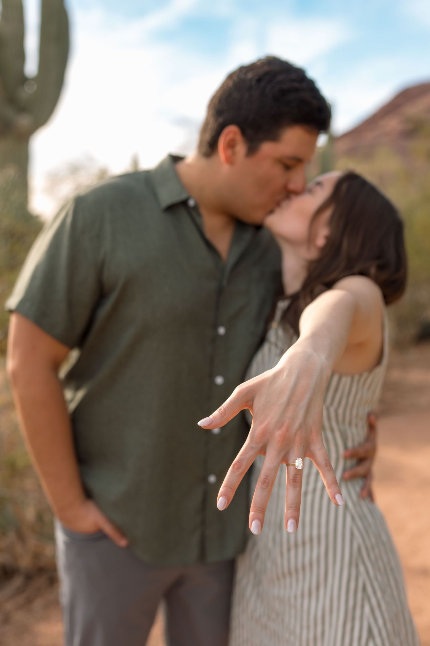 An engaged couple kissing while the woman shows off her diamond engagement ring in a desert setting.