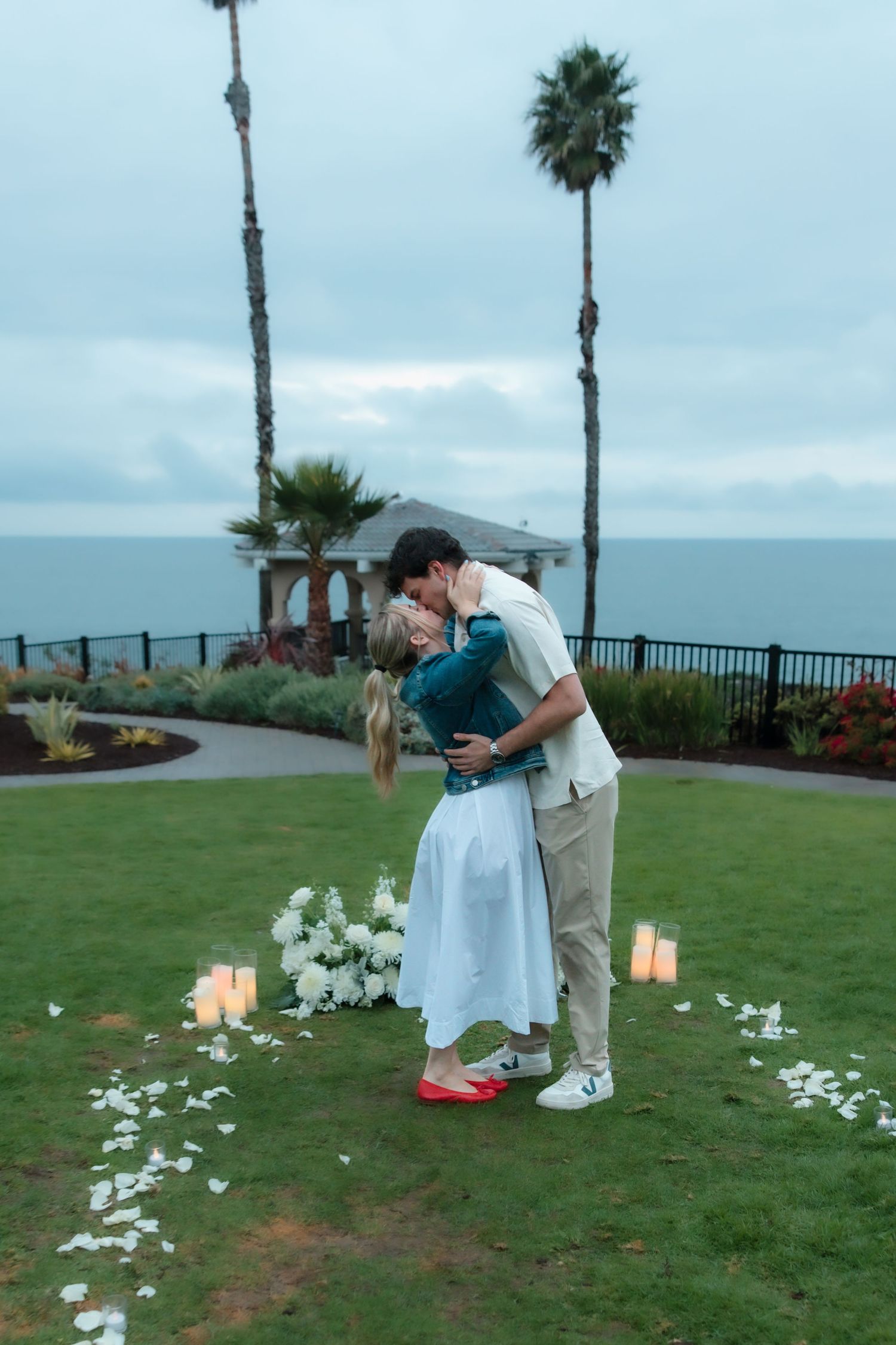 Couple sharing a passionate kiss in a romantic proposal setup with flowers, candles, and ocean view.