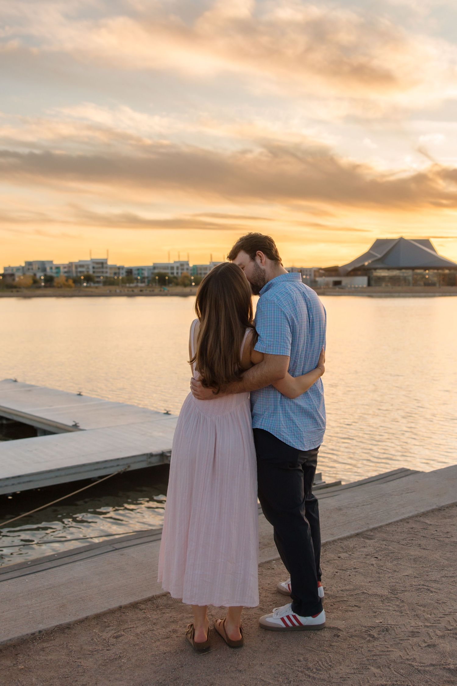 Couple sharing romantic embrace while watching golden sunset over calm lake by dock.