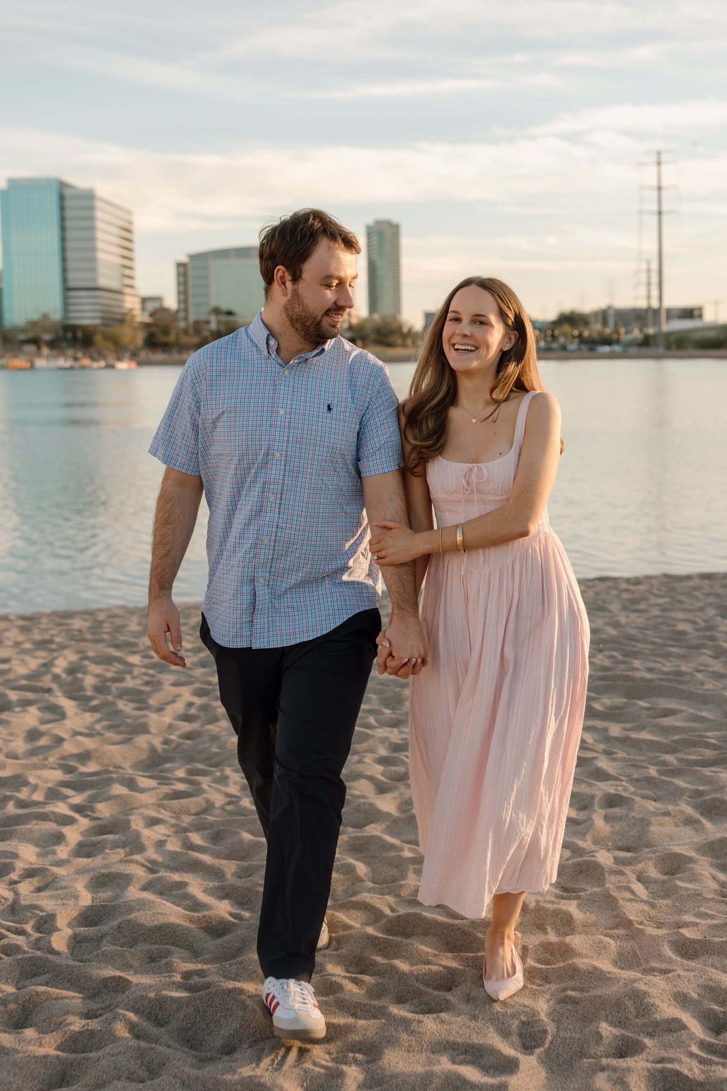 A couple walks hand-in-hand on a sandy beach at sunset, smiling, with a city skyline and water in the background.