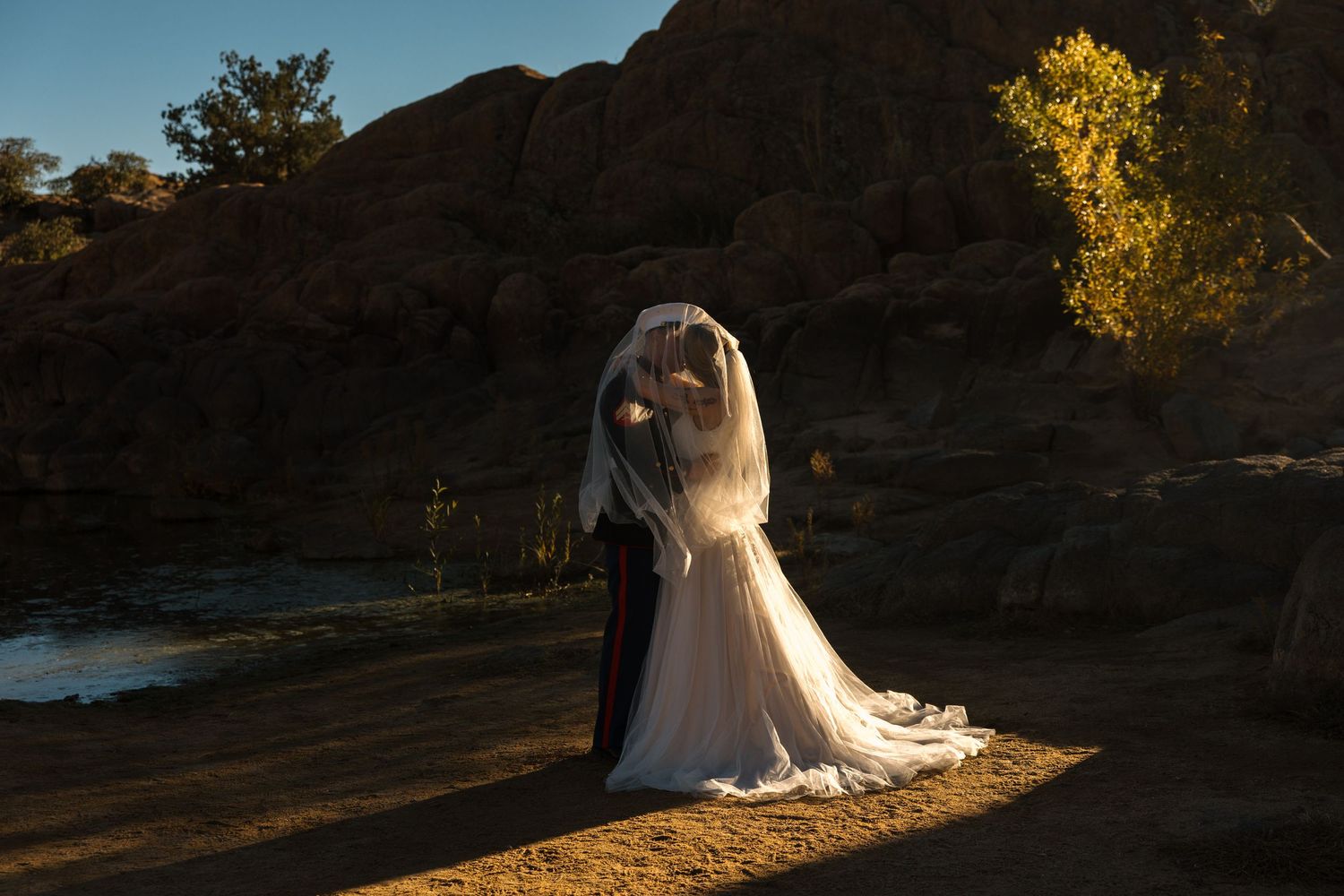 Bride and Marine groom embrace under veil in dramatic golden hour light by lake.