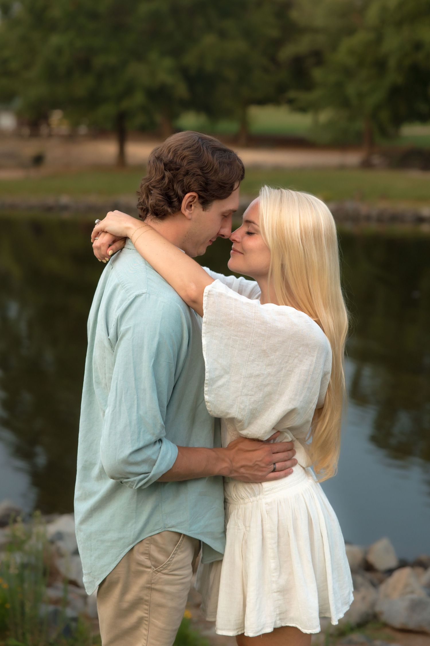 Couple embracing and about to kiss near a peaceful lake surrounded by lush green trees.