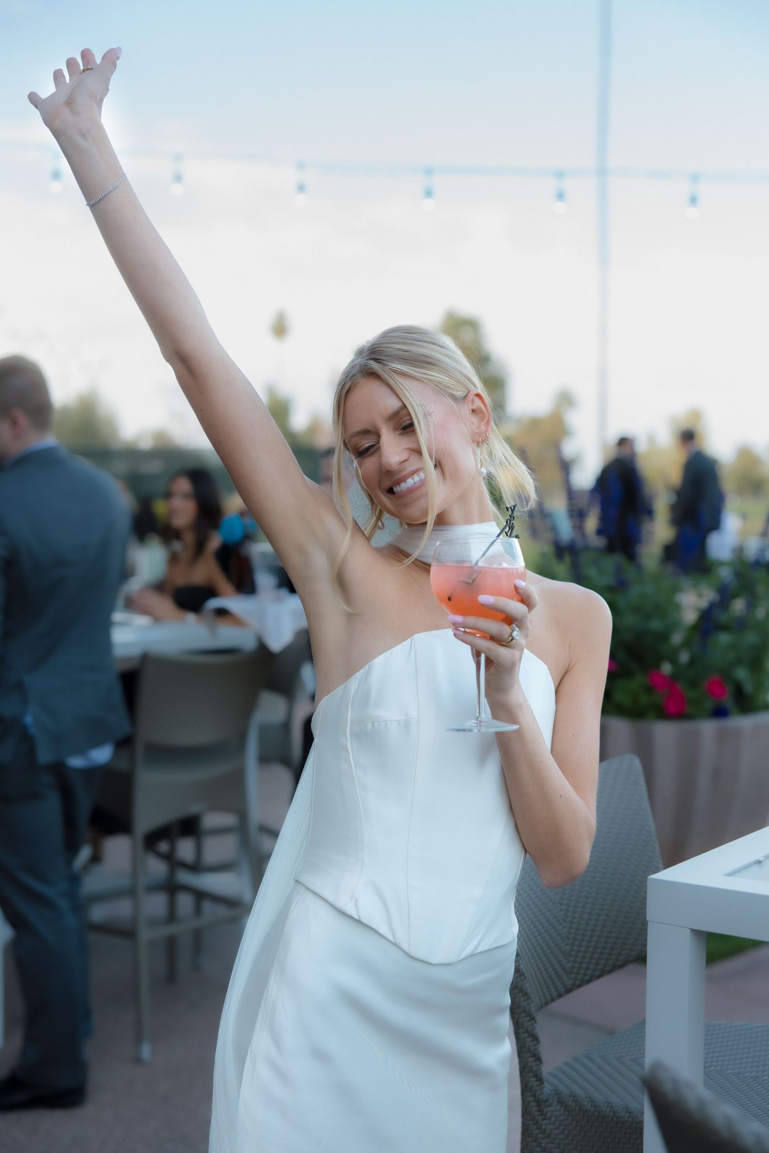Radiant bride in strapless white dress raises arm while holding cocktail at outdoor wedding reception.
