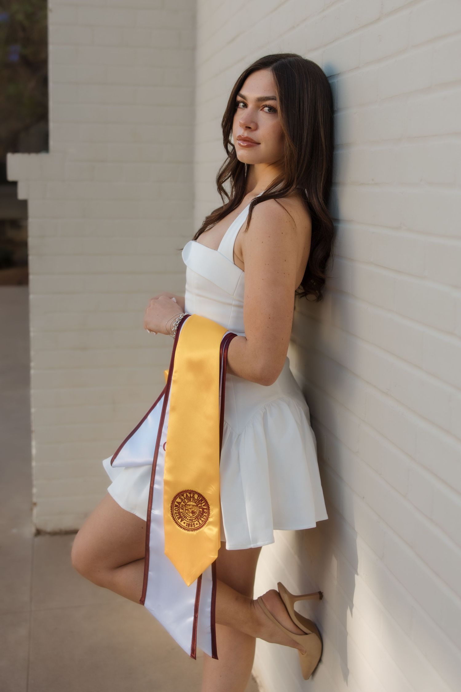 Young woman in white dress holding gold graduation stole leans against white brick wall for graduation photos.
