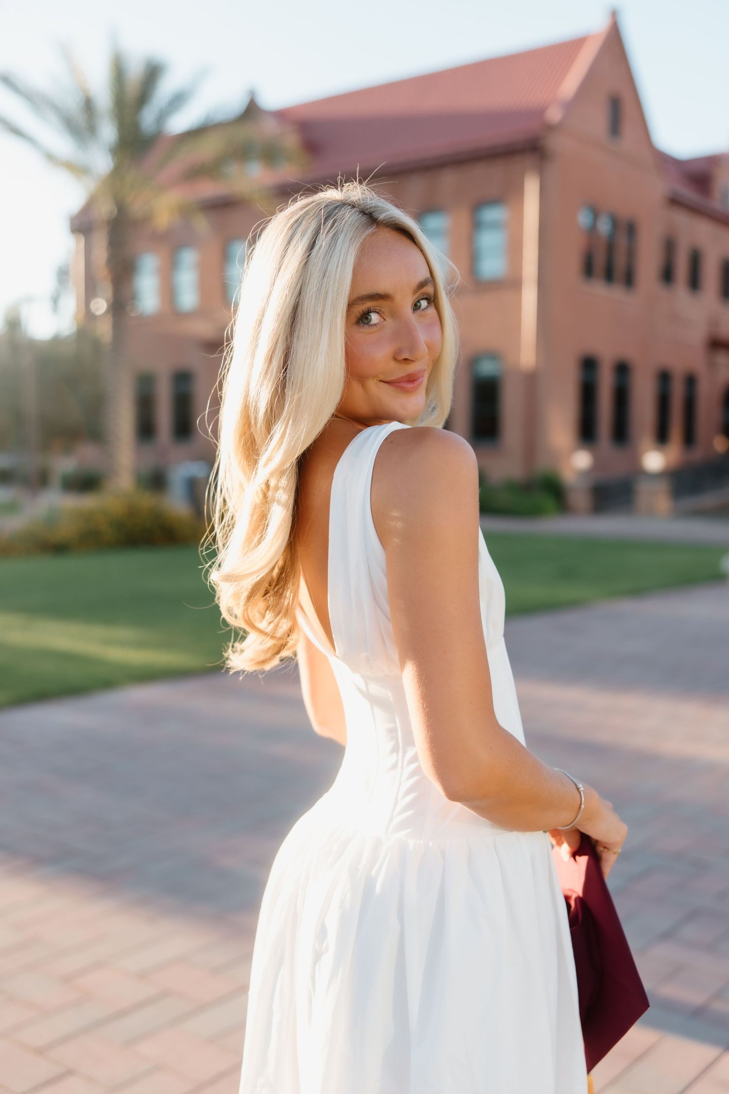 Blonde woman in white backless dress holding burgundy clutch, posing outdoors near brick building on sunny day.