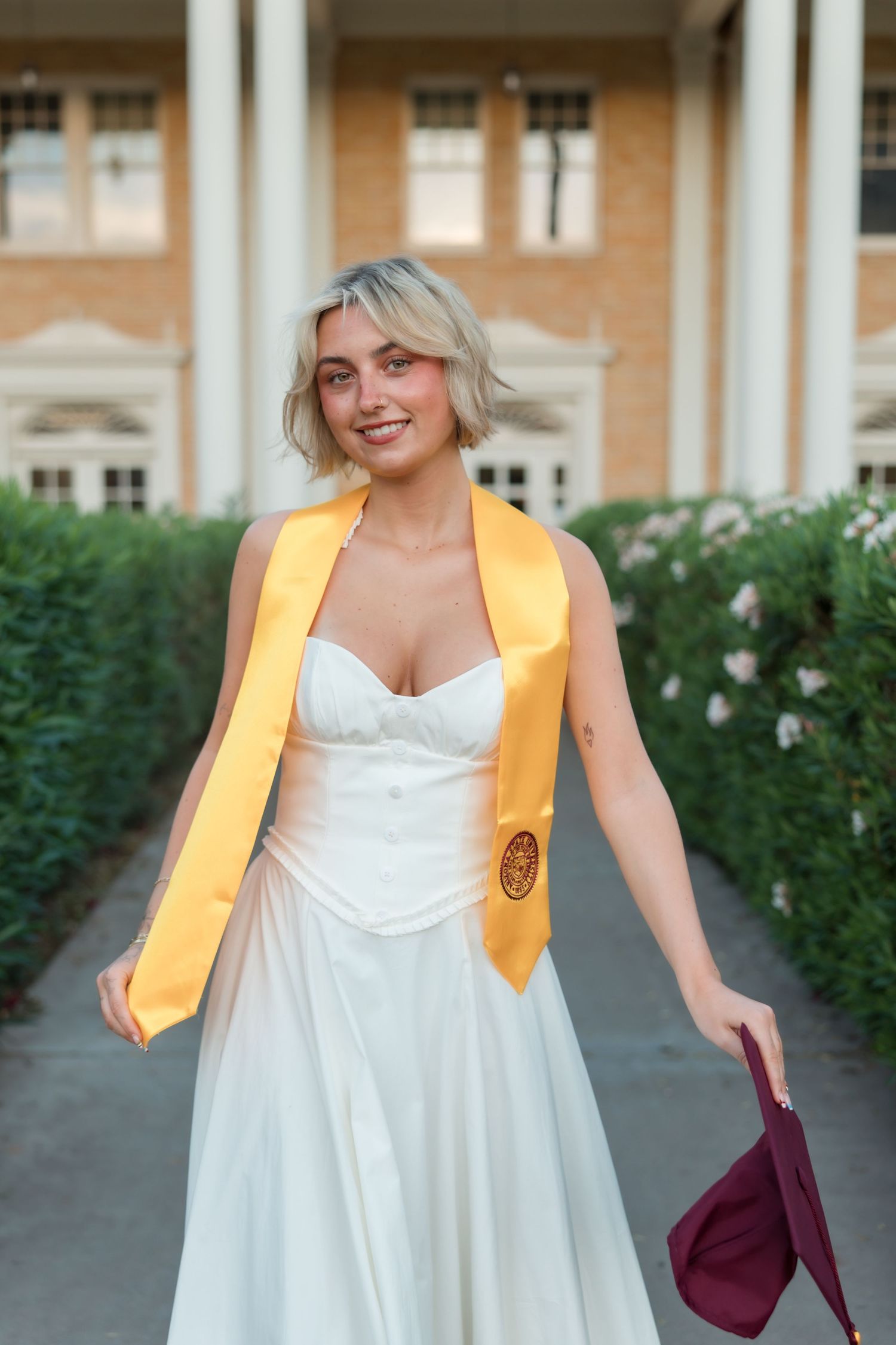 Smiling graduate in white dress and gold honor stole holds maroon cap outdoors near flowering hedges.