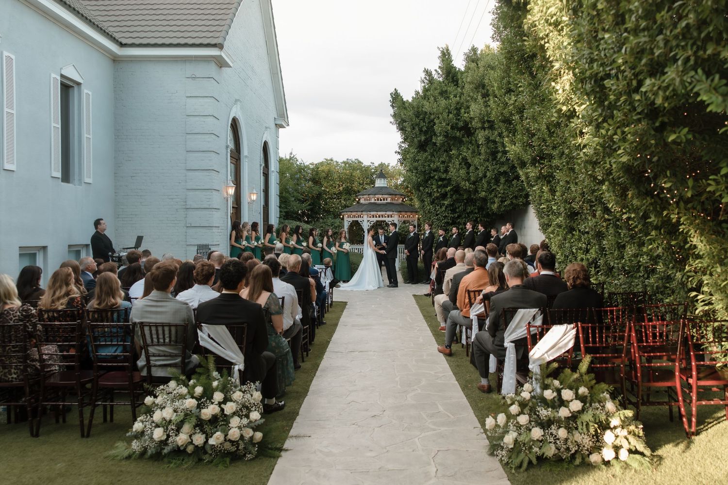 Elegant outdoor wedding ceremony with white aisle runner flanked by floral arrangements and guests.