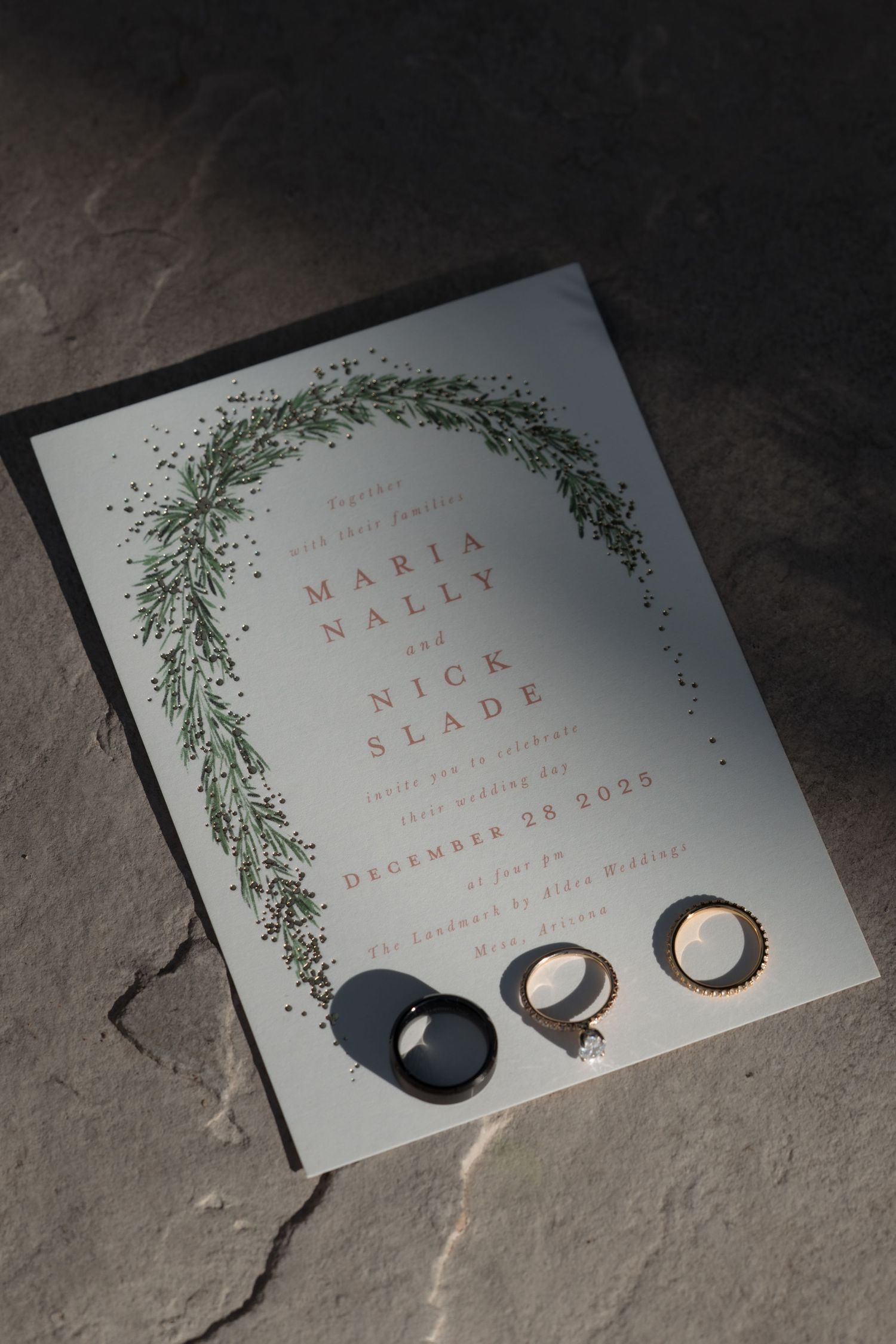 Maria Nally and Nick Slade wedding invitation with greenery wreath and gold rings on stone.