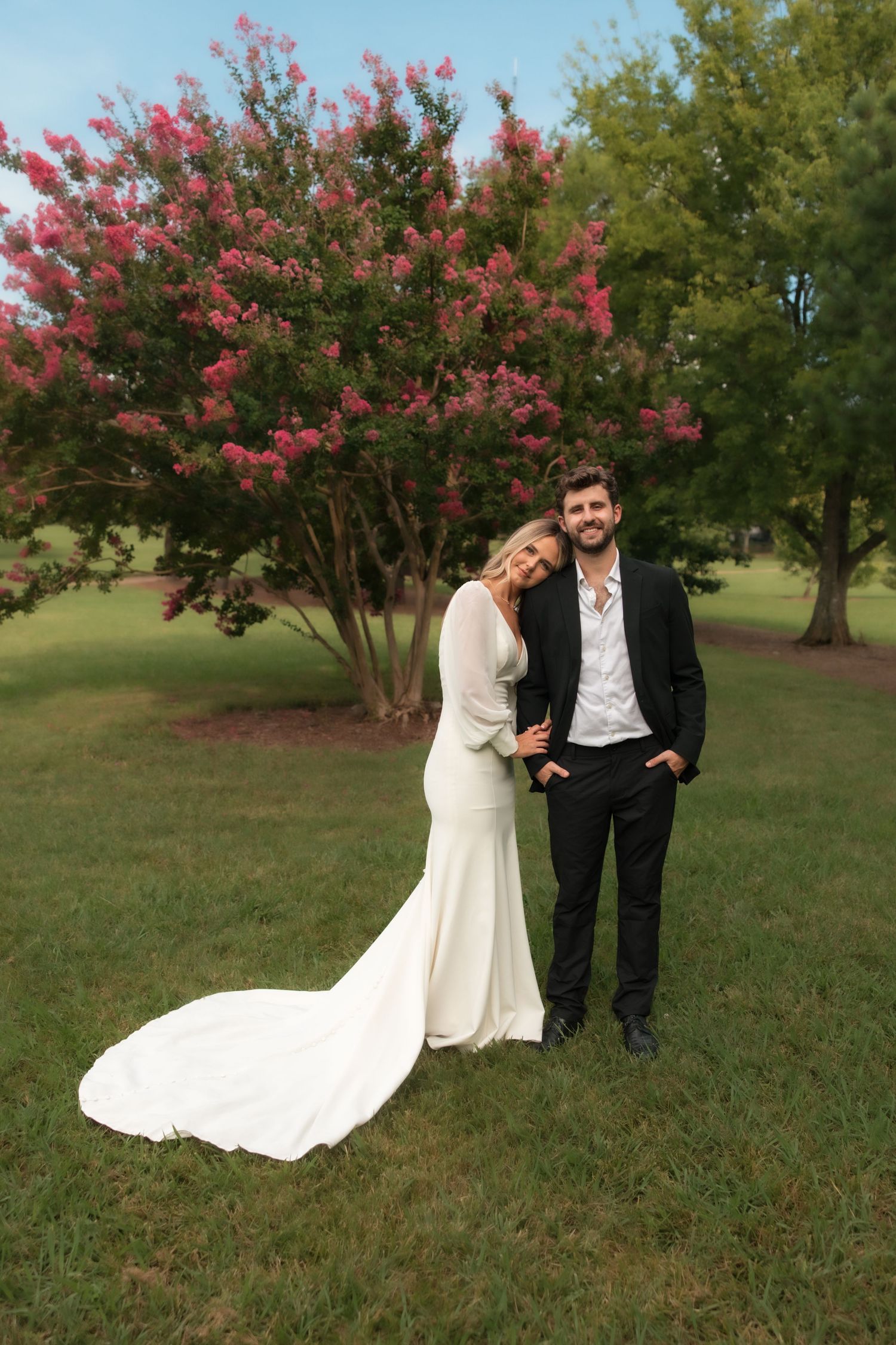 Smiling wedding couple stands before blooming pink crepe myrtle tree, bride in white gown with train.