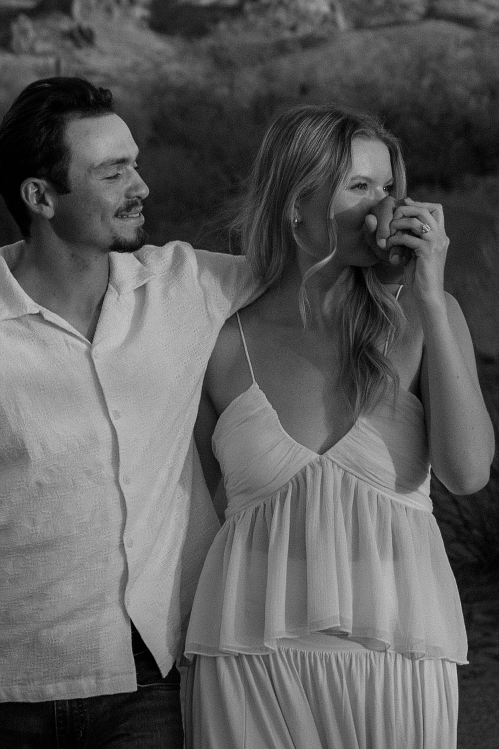 Black and white photo of a smiling couple outdoors, woman in white dress sipping a drink, man in white shirt beside her.