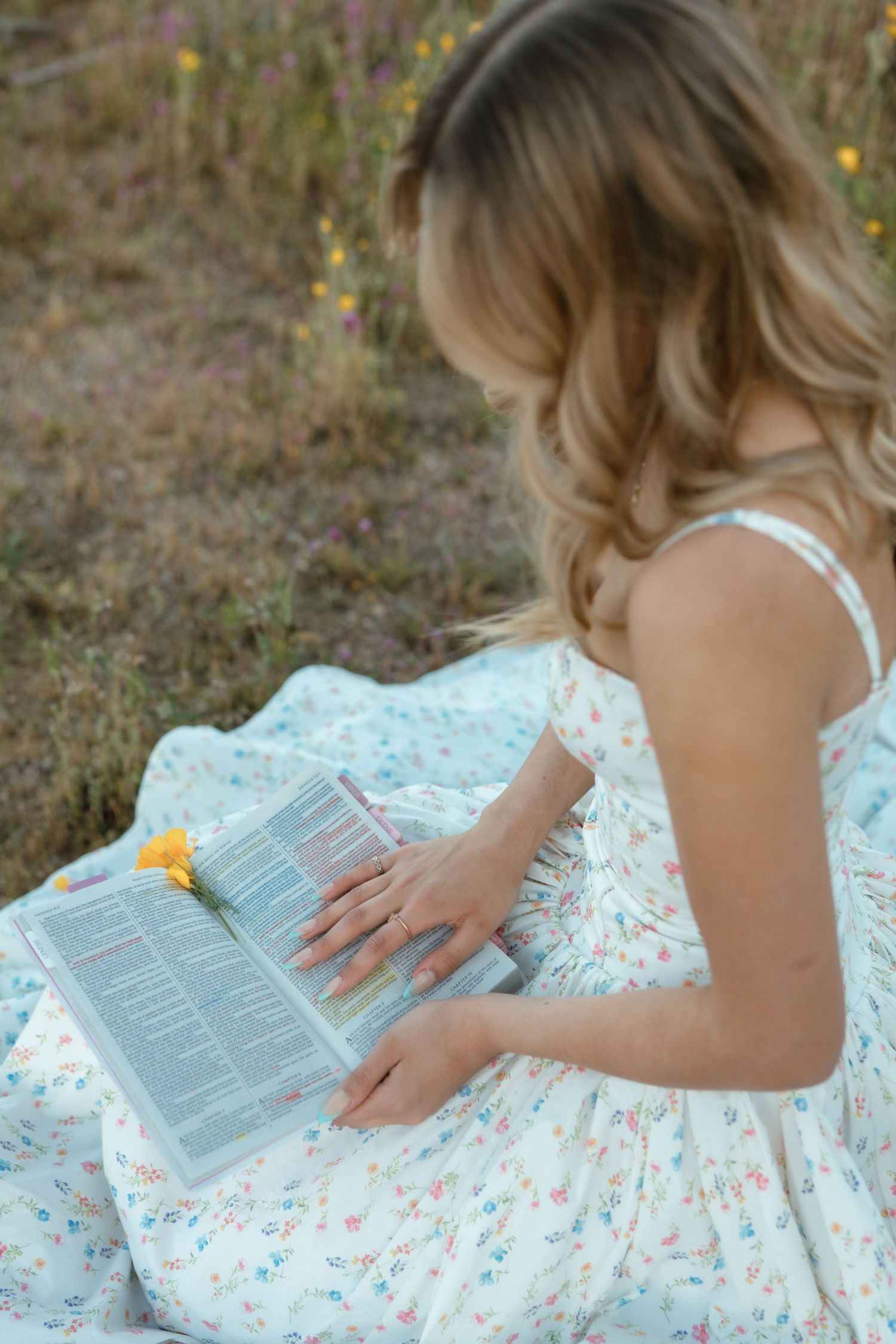 Young woman in floral dress reading a book with a yellow flower outdoors in a wildflower meadow.