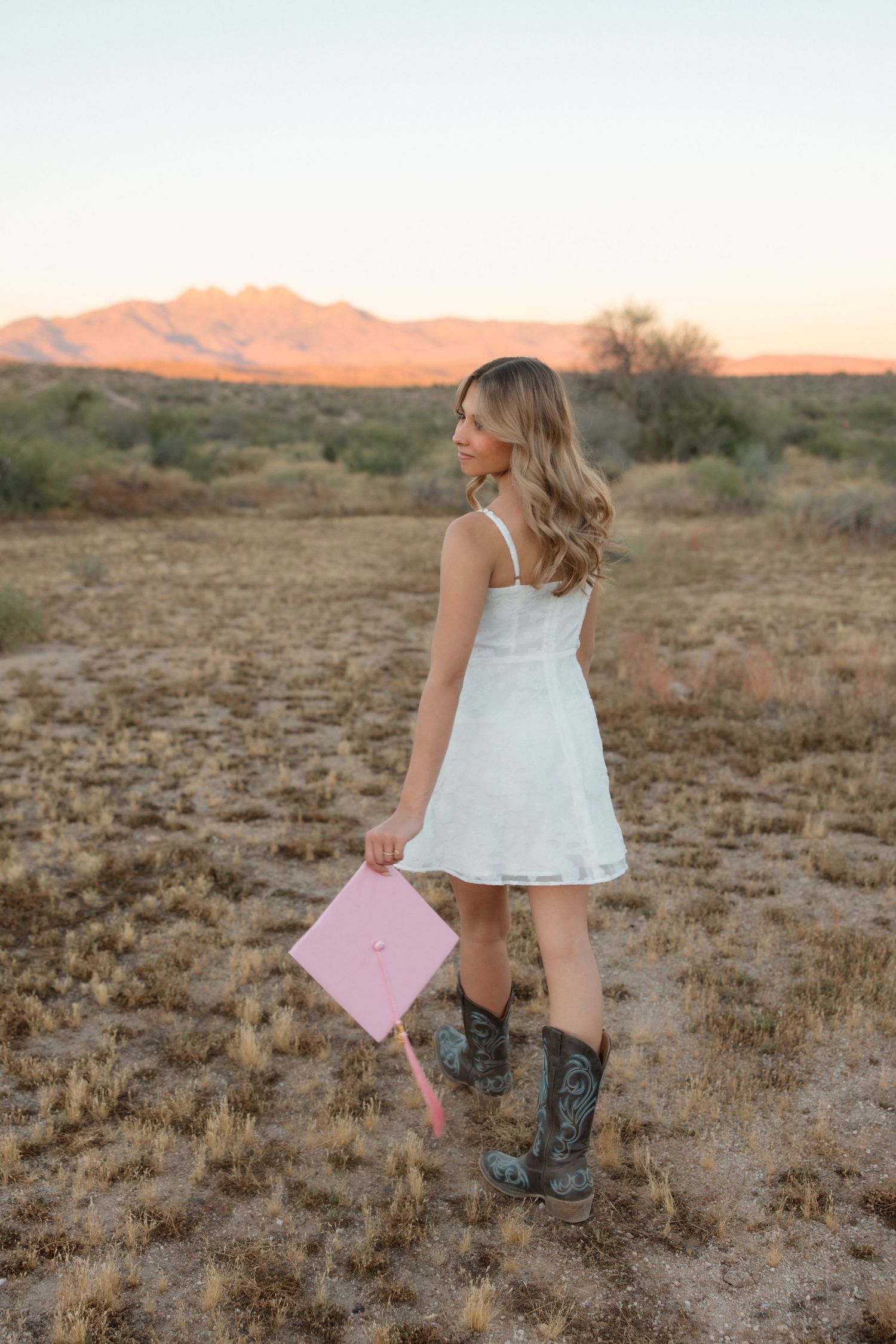 Young woman in white dress and cowboy boots holding pink graduation cap in desert landscape at sunset.