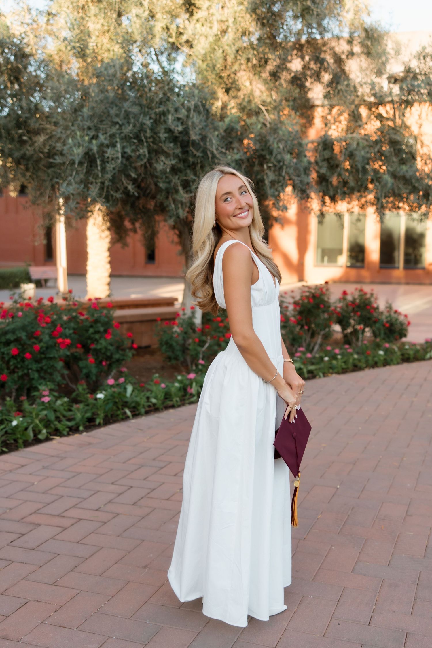 Smiling blonde woman in white dress holding maroon graduation cap outdoors near red roses and brick building.