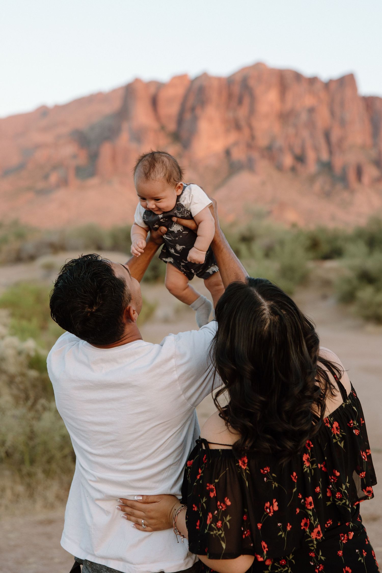 A couple lifts their baby outdoors with red rock mountains in the Arizona desert background at golden hour.