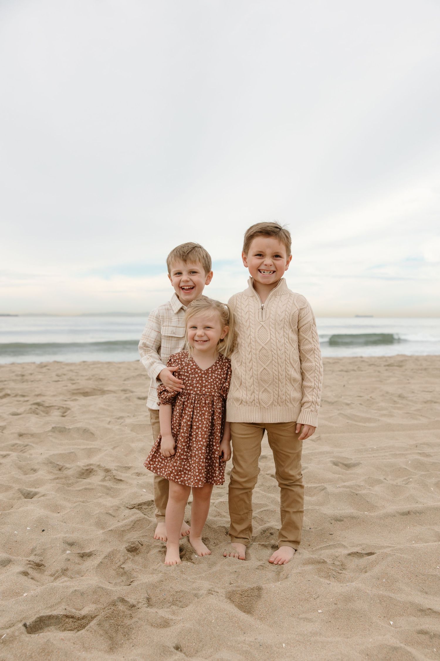 Three young children smiling together on a sandy beach, dressed in warm neutral tones with ocean waves in the background.