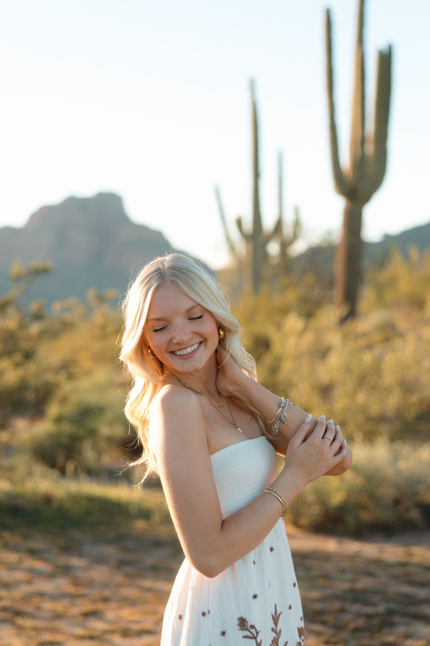 Smiling blonde woman in white strapless dress poses in Arizona desert with saguaro cactus and mountain backdrop.
