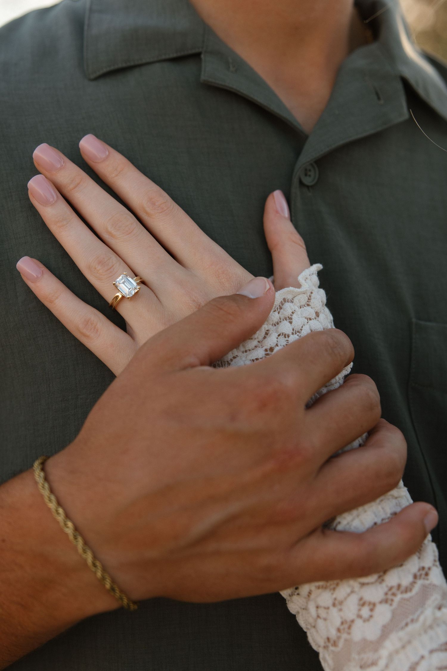 Close-up of an engagement ring on a woman's hand resting on a man's chest, showcasing a stunning emerald-cut diamond.