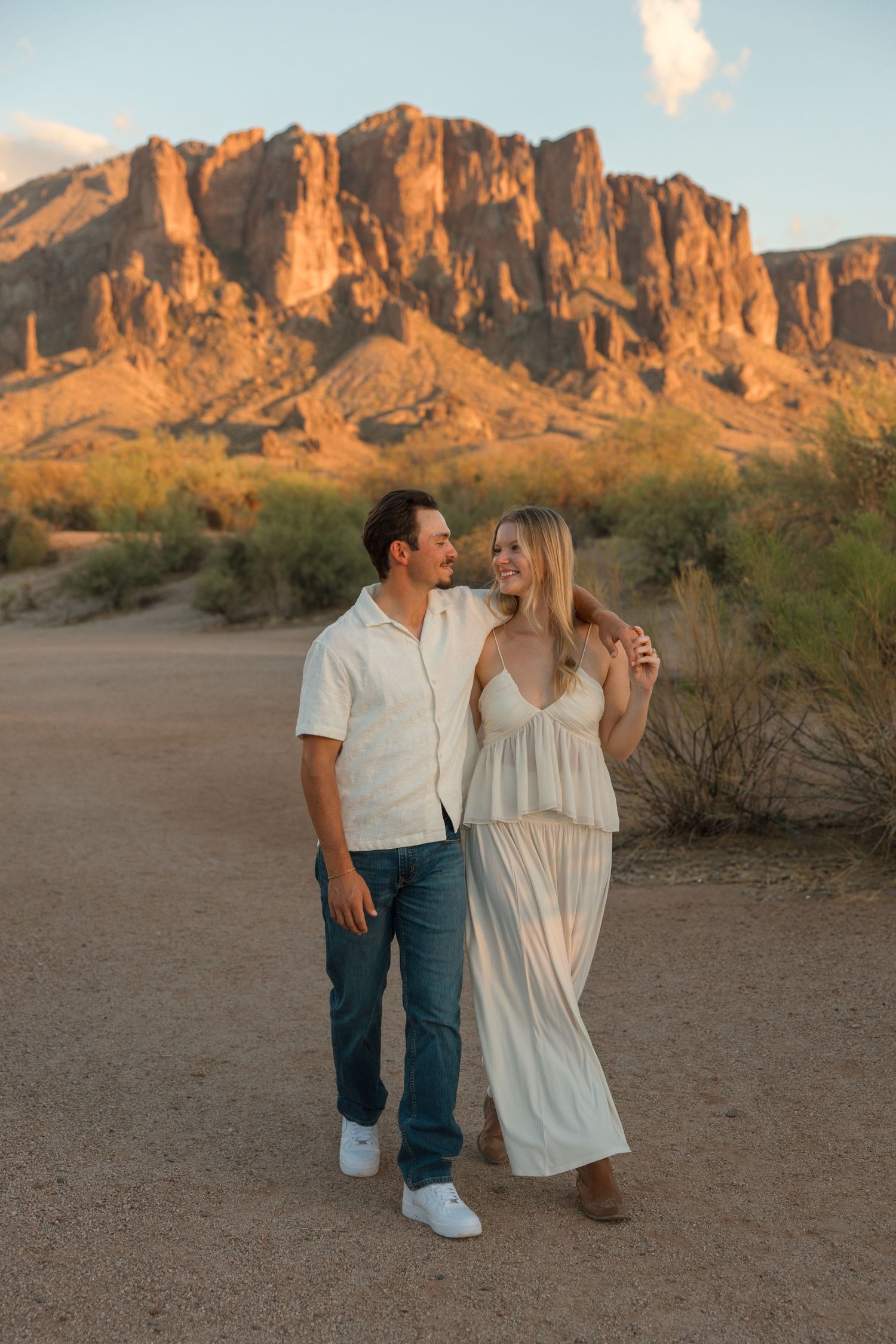 Couple posing in desert landscape with Superstition Mountains at golden hour, wearing white outfits in Arizona.