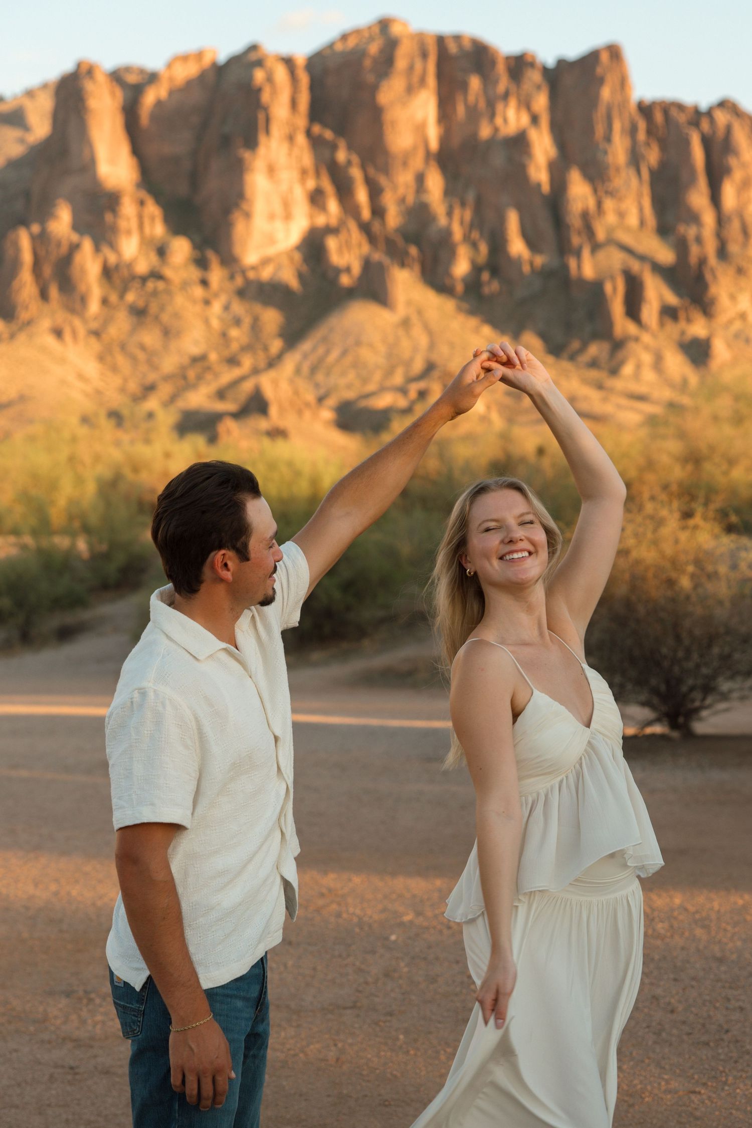 A couple dances outdoors at sunset near Superstition Mountains, Arizona, wearing white outfits.