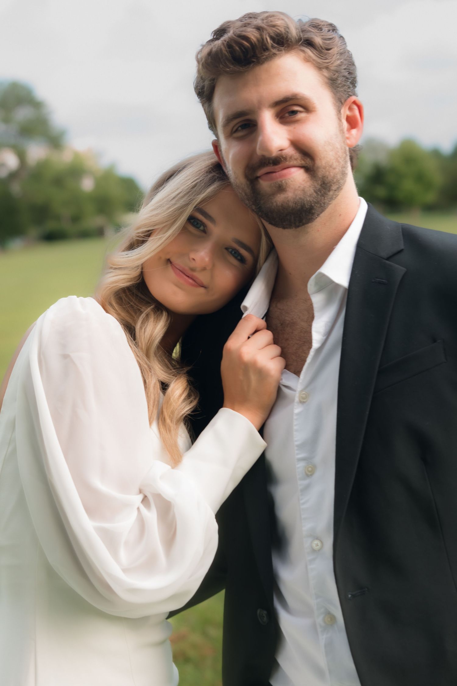 A smiling couple poses outdoors, woman in white dress leaning on bearded man in black suit jacket.