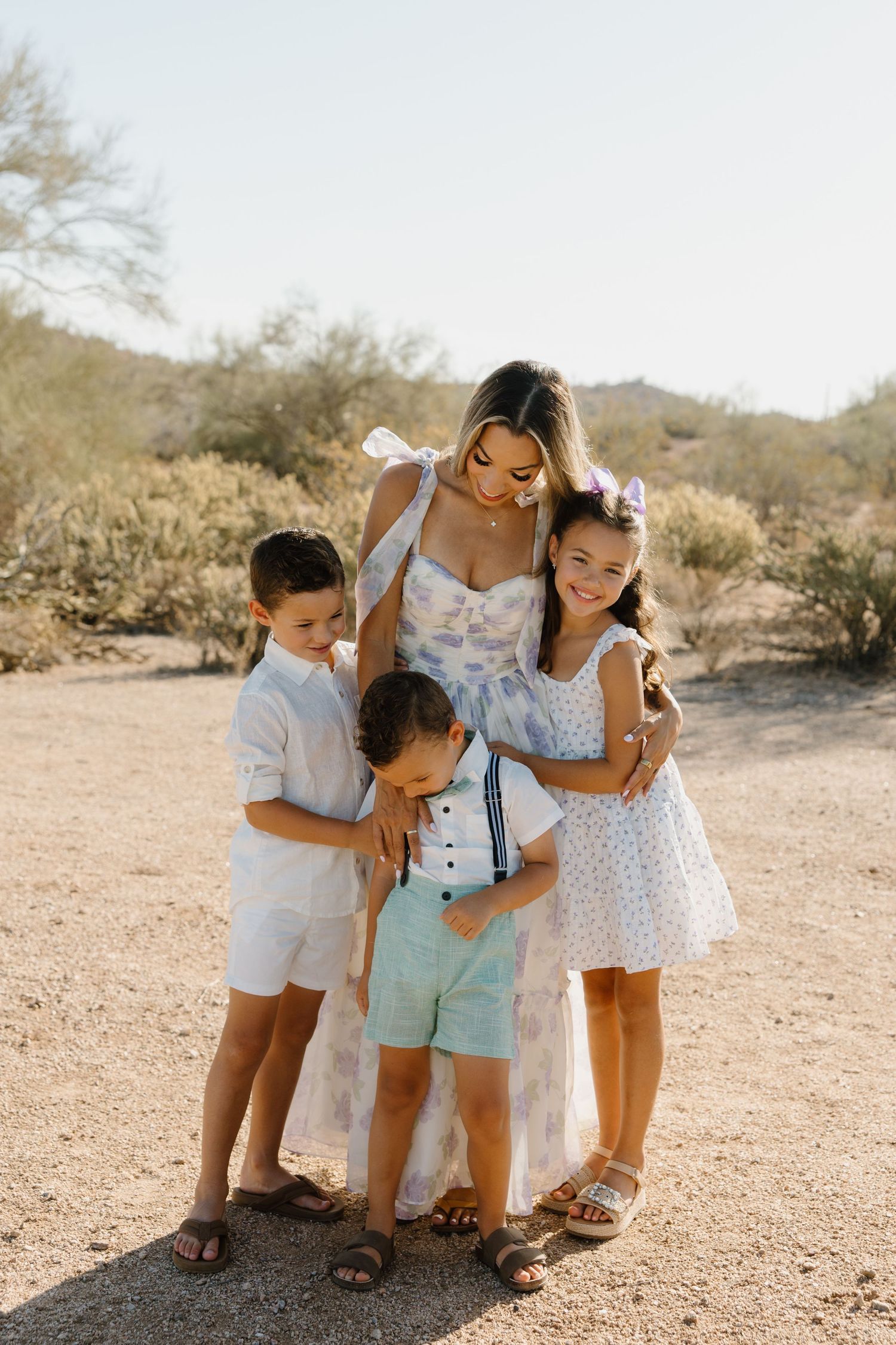 A mother in a floral dress embraces her three young children in a sunny desert landscape.