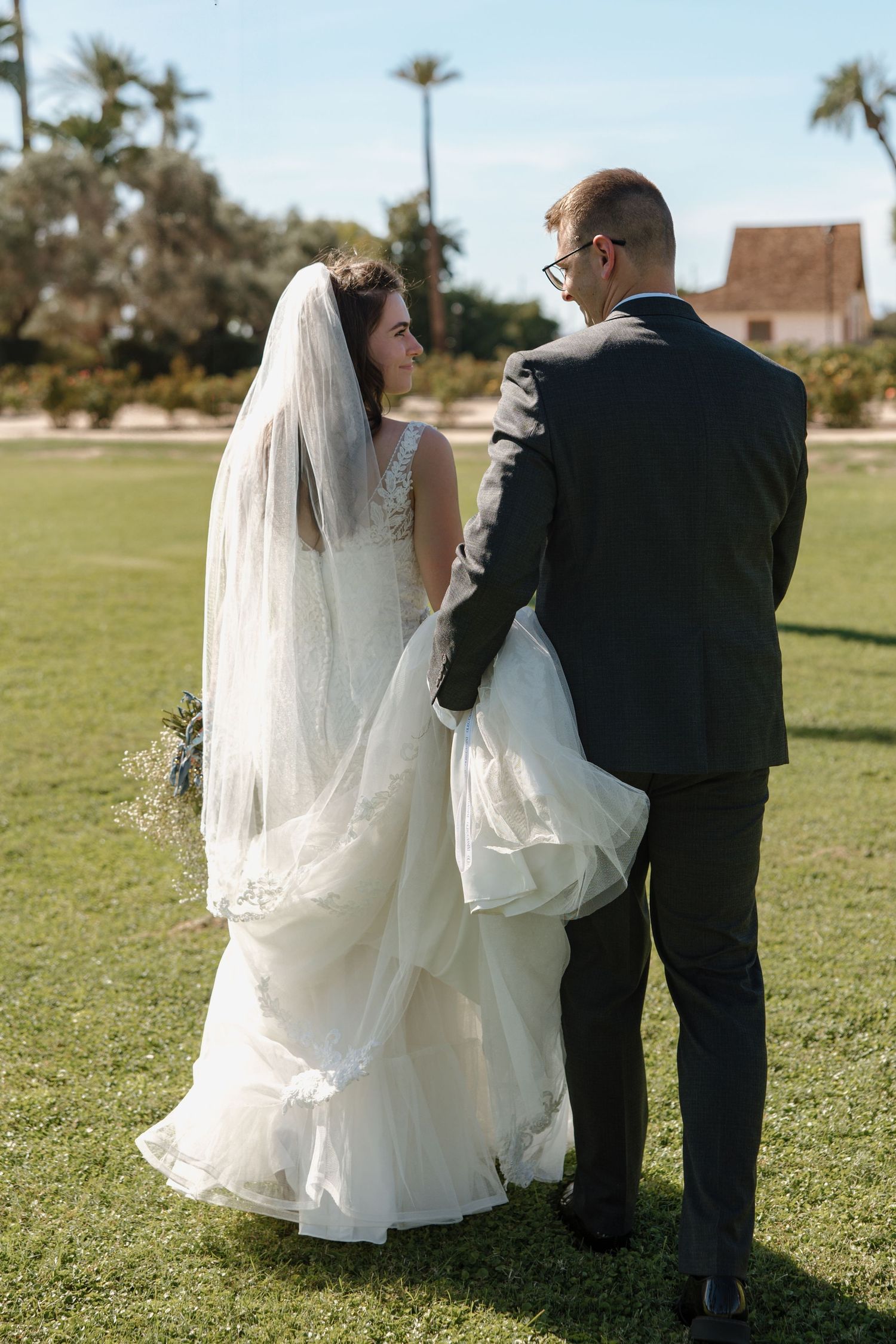 Bride and groom walk hand-in-hand across a green lawn, groom carrying the wedding dress train on sunny day.