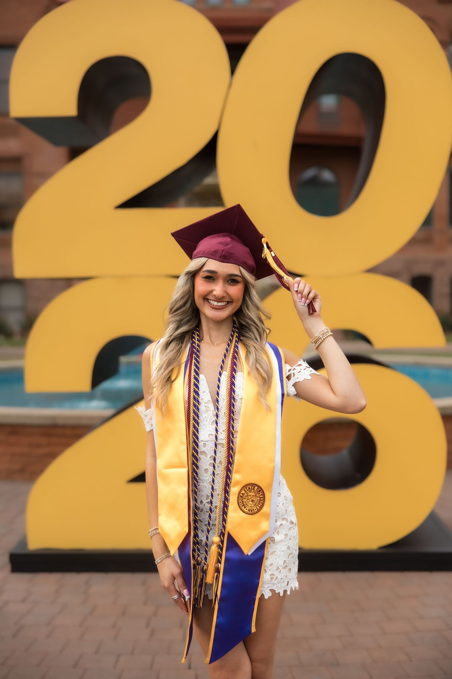 Smiling female graduate in maroon cap and gold stole poses in front of large 2020 graduation sign outdoors.