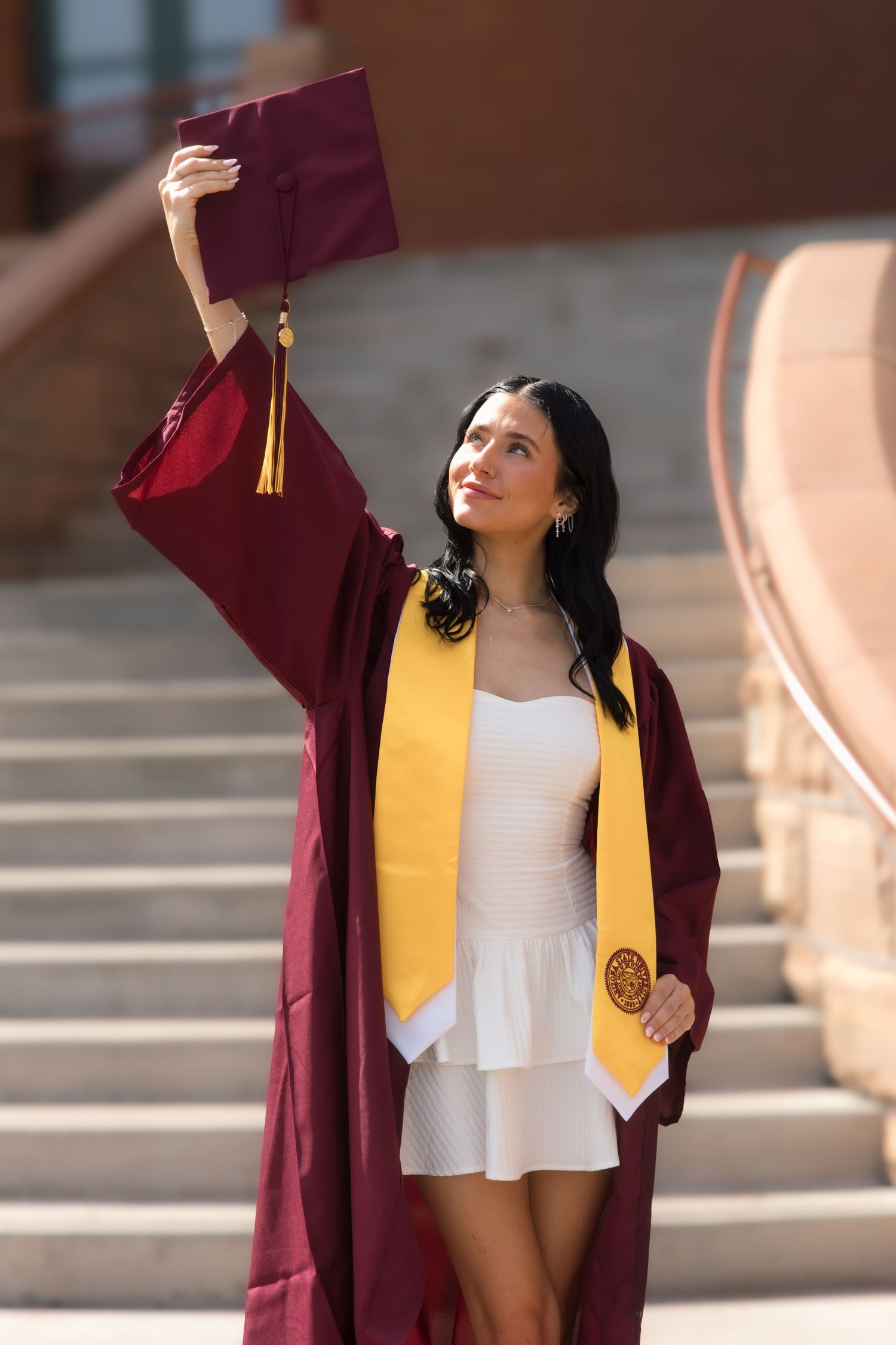 A young woman in a maroon graduation gown and gold stole raises her cap triumphantly on a sunny campus.