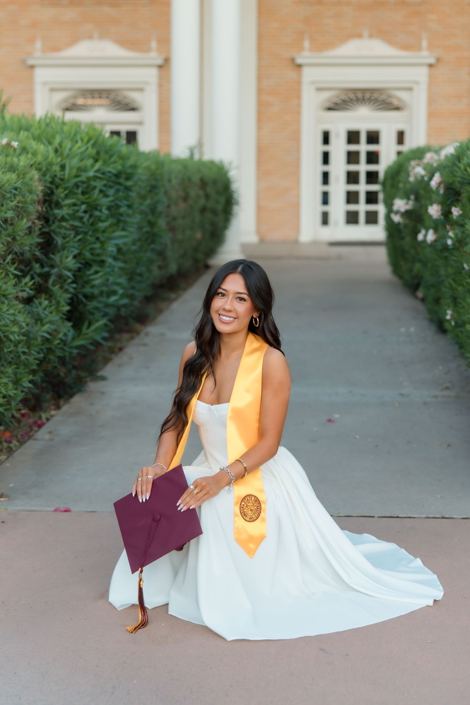 Young woman in white dress and gold graduation stole kneels proudly holding maroon cap outside university building.