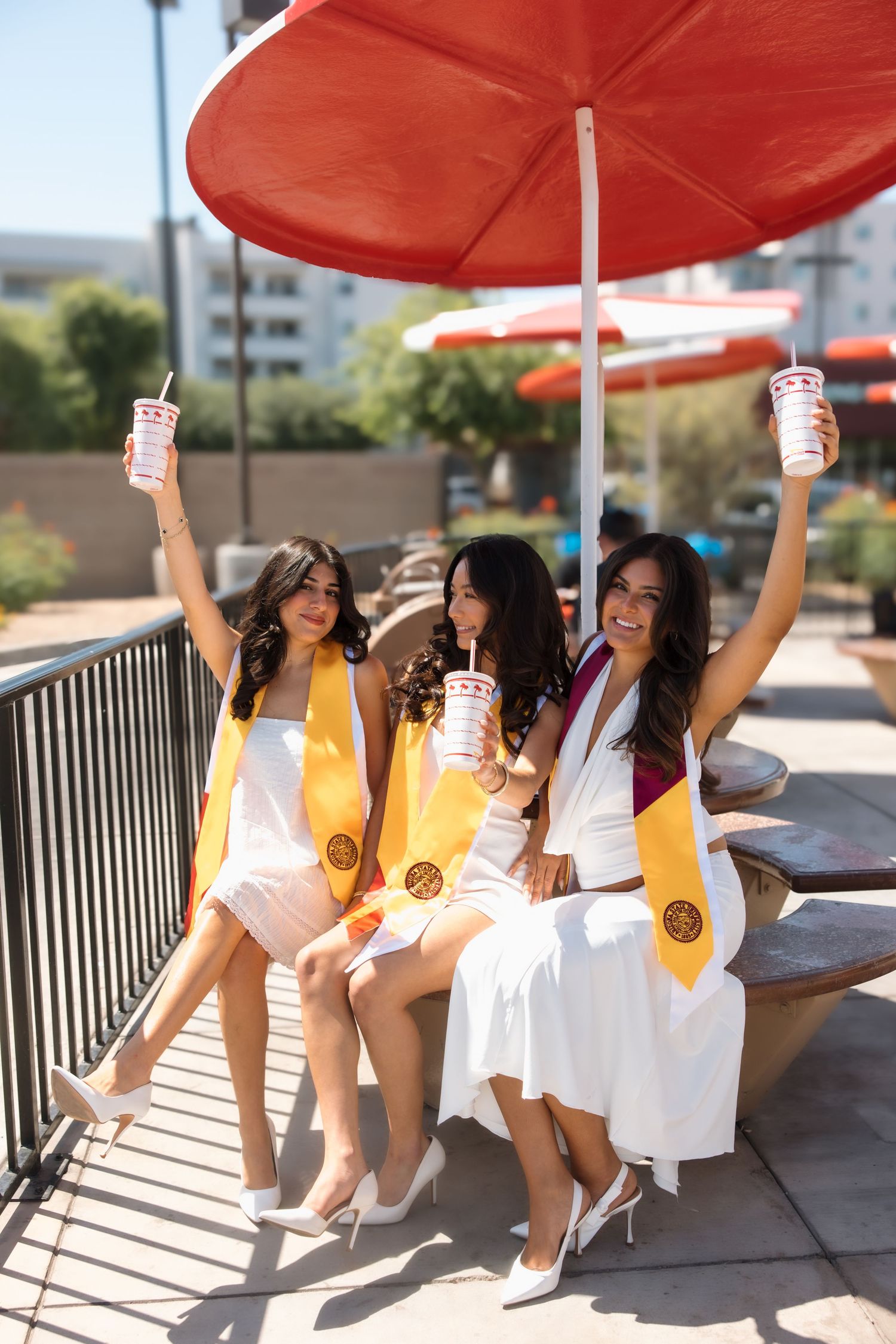 Three college graduates in white dresses and gold stoles raise In-N-Out cups at an outdoor patio table.