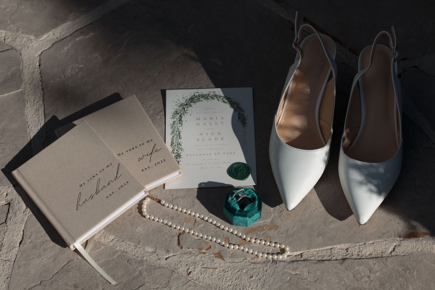 Wedding flat lay featuring white heels, pearl necklace, invitation, and ring box on stone.