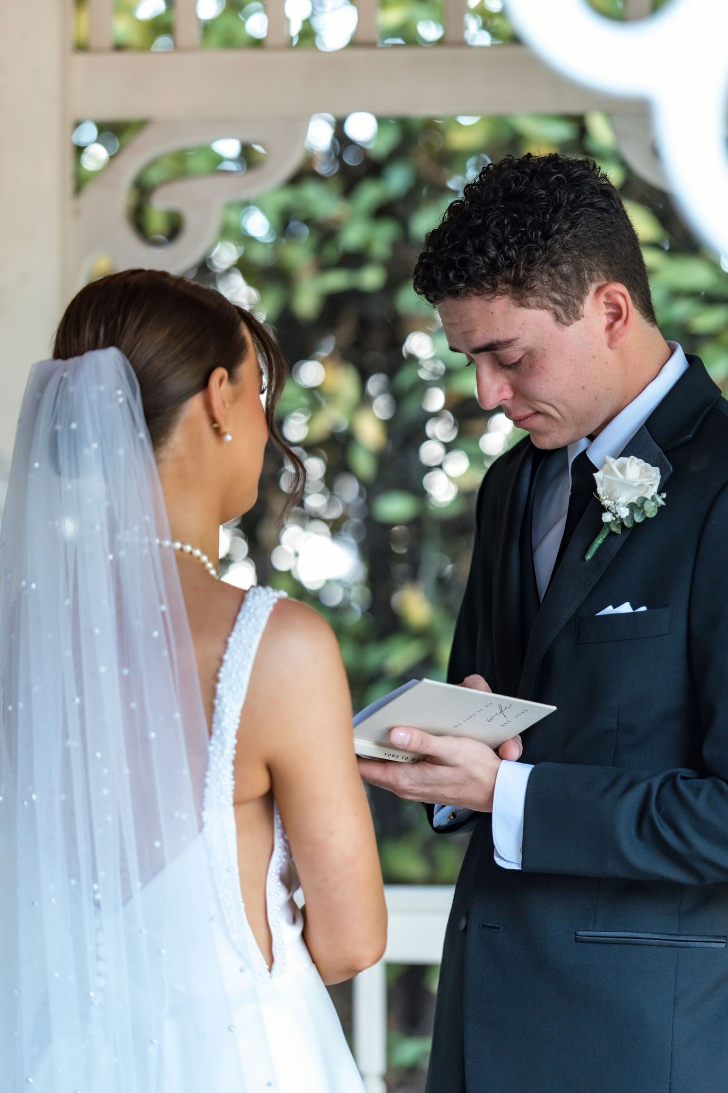Groom reads wedding vows from a book while bride in pearl veil listens at white garden gazebo.